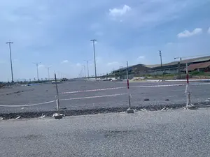 Đây là hình ảnh một công trường xây dựng một con đường lớn hoặc một khu đô thị mới. Mặt đường chưa được trải nhựa mà được phủ một lớp đá dăm. Hai bên đường là hai hàng cột đèn chiếu sáng công cộng mới được lắp đặt, chạy dài về phía xa. Ở phía trước có một hàng rào tạm được dựng lên bằng các cọc cắm trong đế bê tông và căng dây cảnh báo sọc đỏ trắng, cho thấy đây là khu vực đang thi công. Phía xa bên phải có các tòa nhà trông giống như nhà xưởng hoặc nhà kho, và một chiếc cần cẩu xây dựng có thể được nhìn thấy ở phía sau. Rải rác bên lề đường có các vật liệu xây dựng như đống cát và gạch lát. Bầu trời trong xanh, có ít mây.。（日本語訳）。これは、大きな道路または新しい都市区を建設している工事現場の写真です。道路の表面はまだアスファルトで舗装されておらず、砕石の層で覆われています。道路の両側には、新しく設置された公共の街灯の柱が2列あり、遠方まで続いています。手前には、コンクリートの土台に差し込まれた杭と、赤と白の縞模様の警告ロープで張られた仮設の柵があり、ここが工事中の区域であることを示しています。右奥には、工場か倉庫のような建物があり、その後ろに建設用のクレーンが見えます。道端には、砂の山や敷石などの建設資材が点在しています。空は青く澄んでおり、雲はほとんどありません。