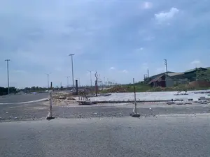 Bức ảnh chụp một công trường xây dựng vào ban ngày, dưới bầu trời có mây. Phía trước là một con đường nhựa. Khu vực chính trong ảnh là một công trình đang thi công, có thể là một khu đô thị mới hoặc một con đường đang được xây dựng. Công trường được rào tạm bằng các cọc gỗ và dây cảnh báo màu đỏ trắng. Bên trong khu vực thi công, có các đống cát và sỏi, cùng với gạch lát vỉa hè đang được thi công dang dở. Nhiều cây xanh non đã được trồng dọc theo vỉa hè và được chống bằng cọc gỗ. Một hàng dài các cột đèn đường mới đã được lắp đặt. Ở phía xa bên phải, có một vài công trình nhà tạm, lán trại và các tòa nhà trông giống nhà xưởng hoặc nhà kho. Ngoài ra còn có các cột điện và đường dây điện. Không có người nào xuất hiện rõ ràng trong khu vực thi công. Không có biển báo hay chữ viết nào có thể đọc được trong ảnh.。（日本語訳）。写真は、曇り空の下、日中の建設現場を撮影したものです。手前にはアスファルトの道路があります。写真の主要なエリアは工事中の現場で、新しい都市区か、建設中の道路である可能性があります。建設現場は、木の杭と赤白の警告テープで仮囲いされています。工事エリア内には、砂と砂利の山があり、それと共に、敷設途中の歩道のタイルがあります。多くの若木が歩道に沿って植えられ、木の杭で支えられています。新しい街灯が長く一列に設置されています。その他にも電柱や電線があります。写真の中には、読み取れる看板や文字はありません。