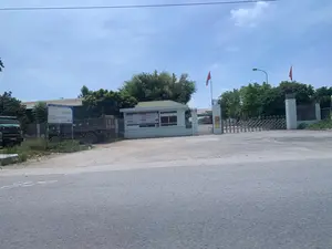 Đây là hình ảnh chụp từ bên ngoài cổng của một công ty, có thể là một nhà máy hoặc xí nghiệp. Cảnh vật cho thấy một con đường nhựa ở phía trước. Lối vào công ty có một cổng sắt xếp màu bạc đang đóng và một nhà bảo vệ màu xanh nhạt. Hai lá cờ đỏ sao vàng của Việt Nam được cắm trên cột. Bên ngoài cổng, phía bên trái, có hai chiếc xe tải đang đỗ và một bảng thông báo lớn màu xanh lam. Trên bảng thông báo có ghi 'CÔNG TY TNHH MAY VÂN HÀ' và 'THÔNG BÁO TUYỂN DỤNG LAO ĐỘNG'. Một bảng hiệu nhỏ hơn màu vàng có ghi tên công ty 'VÂN HÀ' cũng được gắn trên cột cổng. Xung quanh có nhiều cây xanh và phía sau là các tòa nhà của nhà máy. Bầu trời trong xanh, có ít mây.。（日本語訳）。これは、ある会社の門の外から撮影された写真で、工場か企業である可能性があります。風景は、手前にアスファルトの道路があることを示しています。会社への入り口には、閉まっている銀色の伸縮式鉄製門と、水色の警備員室があります。2本のベトナムの金星紅旗がポールに掲げられています。門の外、左側には、2台のトラックが駐車しており、大きな青い掲示板があります。掲示板には「CÔNG TY TNHH MAY VÂN HÀ」（ヴァンハ縫製有限会社）と「THÔNG BÁO TUYỂN DỤNG LAO ĐỘNG」（労働者募集のお知らせ）と書かれています。会社名「VÂN HÀ」（ヴァンハ）と書かれた、より小さな黄色の看板も門の柱に取り付けられています。周囲には多くの緑の木々があり、後ろには工場の建物があります。空は青く澄んでおり、雲はほとんどありません。