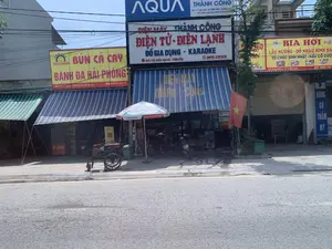 Bối cảnh là một con phố với nhiều cửa hàng kinh doanh san sát nhau tại huyện Thiệu Hóa, tỉnh Thanh Hóa. Con đường nhựa phía trước khá vắng vẻ. Các chi tiết quan sát được bao gồm:。1.  **Bảng hiệu và thông tin chữ viết:**。*   **Quán ăn bên trái:** Biển hiệu màu vàng với dòng chữ 'BÚN CÁ CAY BÁNH ĐA HẢI PHÒNG'. Địa chỉ: 'Tiểu Khu 2 - TT. Thiệu Hóa - Thiệu Hóa - Thanh Hóa'. Số điện thoại: '038 306 97 95 - 039 479 1786'.。*   **Cửa hàng ở giữa:** Cửa hàng 'ĐIỆN MÁY THÀNH CÔNG', kinh doanh 'ĐIỆN TỬ - ĐIỆN LẠNH, ĐỒ GIA DỤNG - KARAOKE'. Địa chỉ: 'Thị trấn Vạn Hà - Thiệu Hóa, Thanh Hóa'. Số điện thoại: '0976 242 909 - 0978 379 973'. Phía trên có biển quảng cáo lớn của hãng 'AQUA'.。*   **Quán ăn bên phải:** Biển hiệu màu vàng quảng cáo 'BIA HƠI', 'LẨU NƯỚNG - ĐỒ NHẬU BÌNH DÂN', và nhận 'TỔ CHỨC SINH NHẬT - NẤU CỖ CƯỚI'. Địa chỉ: 'KHU PHỐ 2 - TT THIỆU HÓA'.。*   **Phòng khám ngoài cùng bên phải:** Biển hiệu dọc màu xanh ghi 'RĂNG SỨ THẨM MỸ'.。2.  **Cảnh vật và hoạt động:**。*   **Loại địa điểm:** Đây là một khu phố thương mại ở một thị trấn.。*   **Vật thể:** Trên vỉa hè có một vài chiếc xe máy, một chiếc xe kéo tay (xe công nông tự chế), một chiếc ô lớn màu trắng xanh của hãng 'Nagakawa' đang mở, và một lá cờ Việt Nam được treo trước cửa hàng điện máy. Các cửa hàng đều có mái hiên sọc xanh.。*   **Hoạt động:** Không có hoạt động mua bán hay con người nào được ghi lại một cách rõ ràng. Khung cảnh thể hiện một ngày bình thường trên một con phố kinh doanh.。（日本語訳）。背景は、タインホア省ティエウホア県にある、多くの商店が隣接する通りです。前方のアスファルトの道はかなり閑散としています。観察される詳細は以下の通りです。1.  **看板と文字情報：**。*   **左側の飲食店：** 黄色い看板に「BÚN CÁ CAY BÁNH ĐA HẢI PHÒNG」（ハイフォン風ピリ辛魚そうめん）の文字。住所：「タインホア省ティエウホア県ティエウホア町第2小区」。電話番号：「038 306 97 95 - 039 479 1786」。*   **中央の店舗：** 「タインコン電器」店、営業品目は「電子機器 - 冷房機器、家庭用品 - カラオケ」。住所：「タインホア省ティエウホア県ヴァンハ町」。電話番号：「0976 242 909 - 0978 379 973」。上部には「AQUA」ブランドの大きな広告看板があります。*   **右側の飲食店：** 黄色い看板で「BIA HƠI」（生ビール）、「LẨU NƯỚNG - ĐỒ NHẬU BÌNH DÂN」（鍋・焼き物 - 大衆向けおつまみ）を広告し、「TỔ CHỨC SINH NHẬT - NẤU CỖ CƯỚI」（誕生日パーティーの開催 - 結婚披露宴の料理）を請け負うとあります。住所：「ティエウホア町第2街区」。*   **一番右端のクリニック：** 青い縦長の看板に「RĂNG SỨ THẨM MỸ」（審美セラミック歯）と書かれています。2.  **風景と活動：**。*   **場所の種類：** これは町にある商店街です。*   **物体：** 歩道には数台のバイク、一台の手押し車（自作の耕運機）、「Nagakawa」ブランドの青と白の大きなパラソルが開かれており、電器店の前にはベトナム国旗が掲げられています。各店舗には青い縞模様の庇があります。*   **活動：** 売買活動や人の姿は、はっきりと記録されていません。この光景は、商店街の平凡な一日を表しています。