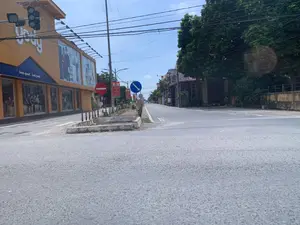 Bối cảnh là một góc phố tại một khu vực đô thị ở Việt Nam vào một ngày nắng. Bên trái là một tòa nhà lớn màu cam, là cửa hàng thời trang của thương hiệu 'YODY'. Trên tường có khẩu hiệu 'look good . feel good'. Bên trong cửa hàng qua lớp kính có thể thấy các ma-nơ-canh đang trưng bày quần áo. Phía trên tòa nhà có hệ thống đèn giao thông. Ở giữa đường có một dải phân cách nhỏ bằng bê tông. Tại đây có cắm một số biển báo giao thông, bao gồm biển 'Cấm đi ngược chiều' (hình tròn đỏ có vạch trắng ngang) và biển chỉ dẫn 'Đi về bên phải' (hình tròn xanh có mũi tên trắng chéo xuống). Ngoài ra còn có các phướn tuyên truyền màu đỏ với dòng chữ 'SỐNG VÀ LÀM VIỆC THEO HIẾN PHÁP VÀ PHÁP LUẬT'. Con đường khá vắng vẻ, chỉ có một chiếc xe máy ở phía xa bên phải. Bên phải đường là dãy nhà dân và nhiều cây xanh lớn. Bầu trời trong xanh và có nhiều dây điện chằng chịt. Khung cảnh chung cho thấy một ngã ba hoặc ngã tư đường phố khá yên tĩnh.。（日本語訳）。背景は、ある晴れた日のベトナムの都市部の一角です。左側は大きなオレンジ色の建物で、「YODY」というブランドのファッションストアです。壁には「look good . feel good」というスローガンがあります。店内ではガラス越しに、服を陳列しているマネキンを見ることができます。建物の上方には信号機があります。道路の中央にはコンクリート製の小さな中央分離帯があります。ここには、「車両進入禁止」（横に白い線が入った赤い円形）の標識と、「右側通行」（白い斜め下向きの矢印がある青い円形）の指示標識を含む、いくつかの交通標識が設置されています。さらに、「憲法と法律に従って生き、働く」という文字が入った赤い宣伝用の垂れ幕もあります。道路はかなり閑散としており、遠く右側にバイクが1台あるだけです。道路の右側には、民家の列とたくさんの大きな緑の木があります。空は青く澄んでおり、たくさんの電線が入り乱れています。全体的な光景は、かなり静かな三叉路または四叉路の通りであることを示しています。