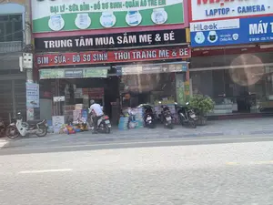 Hình ảnh chụp một dãy cửa hàng mặt tiền trên một con phố. Nổi bật là một cửa hàng lớn tên 'Cường Nguyệt', là một trung tâm mua sắm dành cho mẹ và bé. Các biển hiệu lớn có ghi: 'ĐẠI LÝ - SỮA BỈM - ĐỒ SƠ SINH - QUẦN ÁO TRẺ EM', 'TRUNG TÂM MUA SẮM MẸ & BÉ', và 'BỈM - SỮA - ĐỒ SƠ SINH THỜI TRANG MẸ & BÉ'. Trên biển hiệu có quảng cáo các nhãn hiệu sữa bột như PediaSure, Ensure, Grow. Phía trước cửa hàng, trên vỉa hè, có nhiều thùng hàng (có thể là bỉm hoặc sữa) và đồ chơi trẻ em như ô tô nhựa được bày ra. Một người đàn ông đội mũ bảo hiểm đang ngồi trên xe máy. Nhiều xe máy khác cũng đang đỗ trên vỉa hè. Bên cạnh là một cửa hàng bán đồ điện tử với biển hiệu 'LAPTOP - CAMERA' và thương hiệu camera an ninh 'HiLook', có ghi địa chỉ 'SN 216 - 220, TT. Thi...'. Một tấm biển nhỏ hơn ở bên trái quảng cáo 'File cá hồi Nauy'.。（日本語訳）。ある通りに面した店舗が並ぶ様子を撮影した画像。目立っているのは「Cường Nguyệt」という名前の大きな店で、母親と赤ちゃんのためのショッピングセンターです。大きな看板には、「代理店 - 粉ミルク・おむつ - 新生児用品 - 子供服」、「ママ＆ベビーショッピングセンター」、そして「おむつ - 粉ミルク - 新生児用品 ママ＆ベビーファッション」と書かれています。看板には、PediaSure、Ensure、Growなどの粉ミルクブランドの広告があります。店の前、歩道上には、多くの商品箱（おむつか粉ミルクの可能性あり）と、プラスチック製の自動車のような子供のおもちゃが陳列されています。ヘルメットをかぶった男性がバイクに座っています。他にも多くのバイクが歩道に停まっています。隣には、「LAPTOP - CAMERA」という看板と防犯カメラブランド「HiLook」を掲げた電化製品店があり、住所「SN 216 - 220, TT. Thi...」が記載されています。左側にある小さな看板には、「ノルウェー産サーモンフィレ」の広告が出ています。