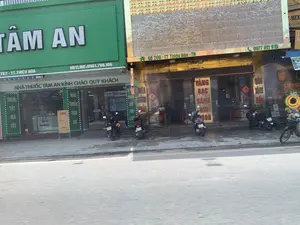 Bức ảnh chụp cảnh một con phố tại Thị trấn Thiệu Hóa, tỉnh Thanh Hóa. Bên trái là một hiệu thuốc có mặt tiền màu xanh lá cây với biển hiệu lớn ghi 'TÂM AN'. Các thông tin khác trên biển hiệu bao gồm 'TK2 - TT. THIỆU HÓA', 'HOTLINE: 0961.798.106', và dòng chữ 'NHÀ THUỐC TÂM AN KÍNH CHÀO QUÝ KHÁCH'. Tại lối vào có các biển dọc ghi 'THUỐC TỐT GIÁ TỐT' và 'NHÀ THUỐC TÂM AN'.。Bên phải là một cửa hàng có mặt tiền màu vàng, dường như là một tiệm vàng bạc. Biển hiệu của cửa hàng này ghi địa chỉ 'SỐ 208 - TT Thiệu Hóa - TH' và số điện thoại '0977 911 915'. Một biển hiệu dọc có thể đọc được chữ 'VÀNG BẠC SÁNH GHI 9999'.。Trên vỉa hè trước các cửa hàng có một vài chiếc xe máy đang đỗ. Con đường ở phía trước là đường nhựa. Khung cảnh cho thấy một ngày hoạt động kinh doanh bình thường trên một con phố ở Việt Nam.。（日本語訳）。タインホア省、ティエウホア町の通りの風景を撮影した写真です。左側には、緑色のファサードを持つ薬局があり、「TÂM AN」と書かれた大きな看板が掲げられています。看板にあるその他の情報には、「TK2 - TT. THIỆU HÓA」、「HOTLINE: 0961.798.106」、そして「NHÀ THUỐC TÂM AN KÍNH CHÀO QUÝ KHÁCH」という文言が含まれています。入口には、「THUỐC TỐT GIÁ TỐT」と「NHÀ THUỐC TÂM AN」と書かれた縦長の看板があります。右側には、黄色のファサードを持つ店があり、金銀店のようです。この店の看板には、「SỐ 208 - TT Thiệu Hóa - TH」という住所と、「0977 911 915」という電話番号が書かれています。縦長の看板には、「VÀNG BẠC SÁNH GHI 9999」という文字が読み取れます。店の前の歩道には、数台のバイクが停まっています。手前の道はアスファルトの道路です。この光景は、ベトナムのある通りの通常の営業日を示しています。