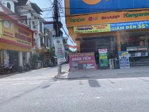 Đây là hình ảnh chụp một góc phố tại một khu đô thị ở Việt Nam, có các cửa hàng kinh doanh san sát nhau. Bên trái là một tiệm vàng bạc tên "VÀNG BẠC SÁNH GHI" với biển hiệu màu vàng, có ghi thêm "TRANG SỨC PHONG THỦY". Bên phải là một cửa hàng điện máy lớn với biển hiệu màu cam và xanh dương, quảng cáo các thương hiệu "oppo", "LG", "SHARP", "Kangaroo". Nhiều xe máy đang đậu trên vỉa hè trước các cửa hàng. Ở trung tâm bức ảnh là một cột điện với hệ thống dây điện và cáp viễn thông chằng chịt. Có rất nhiều biển hiệu và băng rôn quảng cáo được treo lên.。Các thông tin văn bản có thể đọc được bao gồm:。- Tiệm vàng bạc: "VÀNG BẠC SÁNH GHI", "TRANG SỨC PHONG THỦY".。- Cửa hàng điện máy: "oppo", "LG", "SHARP", "Kangaroo".。- Băng rôn quảng cáo điện thoại và TV: "DẦN ĐẦU SMARTPHONE ANDROID", "150 Model - Trả trước từ 10%", "Tivi - Loa Karaoke trả góp 0% GIẢM ĐẾN 35%".。- Băng rôn treo trên cột điện: "Cup Báo Thanh Hóa LẦN THỨ I", "TÀI TRỢ VÀNG VietinBank".。- Biển quảng cáo trên vỉa hè: "BAO GIÁ THỊ TRƯỜDNG HOÀN TIỀN NẾU Ở ĐÂU RẺ HƠN", quảng cáo bếp điện từ Kangaroo giảm giá còn 990.000, và quảng cáo máy lọc nước với các chương trình khuyến mãi như "MIỄN PHÍ lõi lọc lần đầu" và "GIẢM 20% lõi lọc lần sau".。- Chữ trên cửa kính: "BÁN TRẢ GÓP".。（日本語訳）。これはベトナムのある都市部の一角を撮影した写真で、商店が隣接して並んでいます。左側には「VÀNG BẠC SÁNH GHI」という名前の金銀店があり、黄色の看板には「TRANG SỨC PHONG THỦY」と追記されています。右側には大きな家電店があり、オレンジと青の看板で「oppo」、「LG」、「SHARP」、「Kangaroo」のブランドを宣伝しています。多くのバイクが店の前の歩道に停まっています。写真の中央には、電線と通信ケーブルが複雑に絡み合った電柱があります。多くの看板や広告バナーが掲げられています。読み取れるテキスト情報には以下が含まれます：。- 金銀店：「VÀNG BẠC SÁNH GHI」、「TRANG SỨC PHONG THỦY」。- 家電店：「oppo」、「LG」、「SHARP」、「Kangaroo」。- 携帯電話とテレビの広告バナー：「DẦN ĐẦU SMARTPHONE ANDROID」（Androidスマートフォンのリーダー）、「150 Model - Trả trước từ 10%」（150モデル - 頭金10%から）、「Tivi - Loa Karaoke trả góp 0% GIẢM ĐẾN 35%」（テレビ - カラオケスピーカー 分割払い金利0% 最大35%割引）。- 電柱に掲げられたバナー：「Cup Báo Thanh Hóa LẦN THỨ I」（タインホア新聞杯 第1回）、「TÀI TRỢ VÀNG VietinBank」（ゴールドスポンサー VietinBank）。- 歩道上の広告看板：「BAO GIÁ THỊ TRƯỜNG HOÀN TIỀN NẾU Ở ĐÂU RẺ HƠN」（市場価格保証 他店がより安い場合は返金）、KangarooのIHクッキングヒーターが990,000ドンに値下げされている広告、そして「MIỄN PHÍ lõi lọc lần đầu」（初回フィルター無料）や「GIẢM 20% lõi lọc lần sau」（次回フィルター20%割引）などのプロモーションがある浄水器の広告。- ガラス戸の文字：「BÁN TRẢ GÓP」（分割払い販売）。