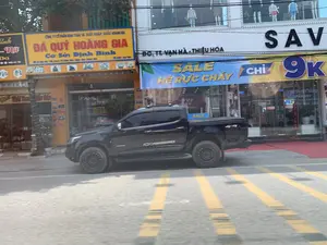 Bản ghi lại các thông tin trên biển hiệu:。1. Biển hiệu màu vàng: 'CÔNG TY CỔ PHẦN KHAI THÁC VÀ XUẤT NHẬP KHẨU HOÀNG GIA', 'ĐÁ QUÝ HOÀNG GIA', 'Cơ Sở: Định Bình'.。2. Biển hiệu màu trắng: 'ĐC, TT. VẠN HÀ - THIỆU HÓA', và tên cửa hàng 'SAV...'.。3. Biểu ngữ màu xanh: 'SALE HÈ RỰC CHÁY', 'CHỈ TỪ 9K'.。4. Các biển hiệu khác có thể đọc được một phần: 'Shop Thời Trang Giầy Dép'.。Mô tả cảnh quan và hoạt động:。Bối cảnh là một con đường ở một khu vực đô thị tại Thị trấn Vạn Hà, huyện Thiệu Hóa. Một chiếc xe bán tải màu đen đang đỗ bên lề đường, phía trước các cửa hàng. Dọc theo con đường là các cửa hàng kinh doanh, bao gồm một cửa hàng đá quý ('ĐÁ QUÝ HOÀNG GIA') và một cửa hàng thời trang lớn. Cửa hàng thời trang đang có chương trình khuyến mãi mùa hè, được quảng cáo bằng biểu ngữ 'SALE HÈ RỰC CHÁY'. Bên trong cửa hàng có các ma-nơ-canh trưng bày quần áo. Mặt đường được trải nhựa và có các vạch kẻ đường màu vàng. Bức ảnh dường như được chụp từ một phương tiện đang di chuyển.。（日本語訳）。看板に記載されている情報の記録：。1. 黄色い看板：「HOANG GIA採掘・輸出入株式会社」、「HOANG GIA宝石」、「支店：ディンビン」。2. 白い看板：「住所、ティエウホア県ヴァンハー町」、および店名「SAV...」。3. 青い横断幕：「燃えるような夏セール」、「わずか9Kから」。4. 部分的に読み取れるその他の看板：「ファッション靴・履物店」。風景と活動の説明：。舞台は、ティエウホア県ヴァンハー町の市街地にある一本の道です。黒いピックアップトラックが、店々の前の路肩に駐車しています。道沿いには、宝石店（「HOANG GIA宝石」）や大きなファッション店を含む商業店舗が並んでいます。ファッション店は夏のプロモーションを実施しており、「燃えるような夏セール」の横断幕で宣伝されています。店内には服を陳列しているマネキンがあります。路面はアスファルトで舗装されており、黄色い車線が引かれています。この写真は移動中の乗り物から撮影されたようです。