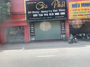 Bức ảnh chụp một con phố với các cửa hàng kinh doanh liền kề tại Thiệu Hóa, Thanh Hóa. Khung cảnh cho thấy một dãy nhà phố thương mại vào ban ngày.。Các thông tin văn bản trên biển hiệu được ghi lại như sau:。1. Cửa hàng ở giữa (biển hiệu màu đen):。- Tên cửa hàng: SPORTS Gia Phát。- Mô tả: Đồ Dùng - Dụng Cụ Thể Thao。- Logo: Nike, Puma, Adidas。- Địa chỉ: Đ/C: SỐ NHÀ 0178 QL 45 TT AN HÀ THIỆU HÓA。- Điện thoại: DD: 090 484 5078。- Biển hiệu dọc: "SN: 0178", "ĐỒ DÙNG THỂ THAO", "CĂNG HÀN VỢT CHO THUÊ BÓNG", "QUẦN ÁO THỂ THAO", "GIẦY THỂ THAO", "IN ÁO ĐỒNG PHỤC".。2. Cửa hàng bên phải (biển hiệu vàng và đỏ):。- Tên cửa hàng: HIỂU MINH。- Mô tả: "THIẾT BỊ TIN HỌC &", "CAMERA GIÁM SÁT", "ĐIỆN NĂNG LƯỢNG", "MÁY TÍNH - LAPTOP - MÁY IN - CA...", "BÁN TRẢ GÓP", "SỬA CHỮA", "THAY THẾ PHỤ..."。- Địa chỉ: CƠ SỞ 1: SN. 174 - TT THIỆU HÓA - THIỆU HÓA。3. Cửa hàng bên trái: Có biển số nhà màu xanh "SỐ NHÀ 180".。Hoạt động và cảnh vật: Cửa hàng thể thao Gia Phát đang đóng cửa, được thể hiện qua cửa cuốn màu xanh lá cây đã kéo xuống. Ngược lại, cửa hàng điện tử Hiểu Minh có vẻ đang mở cửa, với một vài chiếc xe máy (xe gắn máy) đậu ở phía trước và bên trong. Một người đang ngồi trên vỉa hè gần đó. Phía trước các cửa hàng là vỉa hè và một con đường nhựa.。（日本語訳）。タインホア省ティエウホア県にある、商店が隣接する通りの写真。日中の商店街の一角が写っている。看板に書かれている文字情報は以下の通りです。1. 中央の店舗（黒い看板）：。- 店舗名：SPORTS Gia Phát（スポーツ・ザーファット）。- 説明：スポーツ用品・用具。- ロゴ：Nike、Puma、Adidas。- 住所：Đ/C: SỐ NHÀ 0178 QL 45 TT AN HÀ THIỆU HÓA（住所：ティエウホア県アンハ町 国道45号線 178番地）。- 電話番号：DD: 090 484 5078（携帯：090 484 5078）。- 縦看板：「SN: 0178（番地：0178）」、「ĐỒ DÙNG THỂ THAO（スポーツ用品）」、「CĂNG HÀN VỢT CHO THUÊ BÓNG（ラケットのガット張り・ボールレンタル）」、「QUẦN ÁO THỂ THAO（スポーツウェア）」、「GIẦY THỂ THAO（スポーツシューズ）」、「IN ÁO ĐỒNG PHỤC（ユニフォームシャツのプリント）」。2. 右側の店舗（黄色と赤の看板）：。- 店舗名：HIỂU MINH（ヒエウミン）。- 説明：「THIẾT BỊ TIN HỌC &（IT機器＆）」、「CAMERA GIÁM SÁT（監視カメラ）」、「ĐIỆN NĂNG LƯỢNG（太陽光発電）」、「MÁY TÍNH - LAPTOP - MÁY IN - CA...（コンピューター - ラップトップ - プリンター - カ...）」、「BÁN TRẢ GÓP（分割払い販売）」、「SỬA CHỮA（修理）」、「THAY THẾ PHỤ...（部品交換...）」。- 住所：CƠ SỞ 1: SN. 174 - TT THIỆU HÓA - THIỆU HÓA（1号店：ティエウホア県ティエウホア町 174番地）。3. 左側の店舗：青い家屋番号プレートに「SỐ NHÀ 180（180番地）」と書かれている。活動と風景：スポーツ用品店「Gia Phát」は閉店しており、緑色のシャッターが下りていることでそれが示されている。対照的に、電器店「Hiểu Minh」は営業しているようで、店の前と中に数台のバイク（原動機付自転車）が停まっている。近くの歩道に一人の人が座っている。店の前には歩道とアスファルトの道路がある。