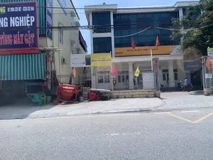 Đây là khung cảnh một con đường tại huyện Thiệu Hóa, tỉnh Thanh Hóa, vào một ngày nắng. Hình ảnh cho thấy một số tòa nhà thương mại. Tòa nhà nổi bật nhất ở bên phải là Bưu điện huyện Thiệu Hóa, một công trình hiện đại có tường kính màu xanh và biển hiệu lớn "BƯU ĐIỆN VIỆT NAM". Một tấm biển khác trên hàng rào ghi rõ đây là "ĐẠI LÝ THU BẢO HIỂM XÃ HỘI TỰ NGUYỆN, BẢO HIỂM Y TẾ" của Bưu điện huyện Thiệu Hóa, và có cả biển hiệu "vinaphone". Bên trong sân bưu điện, có một người đàn ông đội mũ bảo hiểm đang đi bộ gần những chiếc xe máy đang đỗ. Liền kề là một điểm giao dịch của ngân hàng LPBank với biển quảng cáo "LIENVIET24H NGÂN HÀNG SỐ CỦA BẠN". Tòa nhà bên trái là một cửa hàng có mái hiên sọc, với biển hiệu lớn màu xanh lam quảng cáo máy móc nông nghiệp: "...NG NGHIỆP... TÙNG MÁY GẶT" cùng số điện thoại "ĐT: 0904 520 067 - 0336 632 868". Trên vỉa hè, có một máy trộn bê tông màu đỏ của "CTY HÒA PHÁT" và một máy phát điện.。（日本語訳）。これは、晴れた日のタインホア省ティエウホア県のある通りの風景です。右側で最も目立つ建物はティエウホア県郵便局で、青いガラスの壁と「BƯU ĐIỆN VIỆT NAM」という大きな看板がある近代的な建物です。柵にある別の看板には、ここがティエウホア県郵便局の「任意社会保険・健康保険徴収代理店」であることが明記されており、「vinaphone」の看板もあります。郵便局の敷地内では、ヘルメットをかぶった男性が駐車されているバイクの近くを歩いています。隣にはLPBankの取引拠点があり、「LIENVIET24H あなたのデジタルバンク」という広告看板があります。左側の建物は縞模様の庇がある店舗で、農業機械を宣伝する大きな青い看板があり、「...NG NGHIỆP... TÙNG MÁY GẶT」という文字と電話番号「ĐT: 0904 520 067 - 0336 632 868」が書かれています。歩道には、「CTY HÒA PHÁT」の赤いコンクリートミキサーと発電機が1台あります。