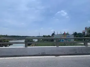 Bức ảnh được chụp từ trên một cây cầu, nhìn ra một con sông rộng. Ở phía trước là lan can bê tông và kim loại của cây cầu. Bên dưới là dòng sông có nước màu phù sa, và bờ sông bên kia có nhiều cây cối xanh tươi. Ở phía xa có thể thấy một cột điện cao thế. Về phía bên phải, có một tòa nhà với một tấm pa-nô tuyên truyền lớn vẽ hình người và lá cờ Đảng Cộng sản Việt Nam (cờ búa liềm). Có nhiều lá cờ phướn với các màu sắc khác nhau đang bay. Chữ viết trên tấm pa-nô không thể đọc rõ. Khung cảnh tổng thể là một ngày nắng, trời có mây, tại một khu vực ven sông ở Việt Nam.。（日本語訳）。写真は橋の上から撮影されたもので、広い川を見渡しています。手前には、橋のコンクリートと金属製の手すりがあります。下には、沖積土色の水が流れる川があり、対岸には緑豊かな木々が多く生い茂っています。遠くには、高圧電線の鉄塔が見えます。右手には、人物とベトナム共産党の旗（鎌と槌の旗）が描かれた大きなプロパガンダの看板がある建物があります。様々な色の多くの幟旗がはためいています。看板の文字ははっきりと読み取れません。全体的な風景は、ベトナムの川沿いの地域で、晴れていて雲がある日です。