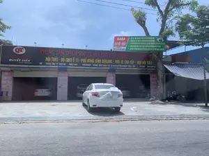Hình ảnh chụp mặt tiền của một trung tâm chăm sóc xe hơi tên là 'Quyết Tiến Auto Detailing'. Đây là một gara ô tô nằm ven đường. Phía trước có một chiếc ô tô màu trắng đang đỗ, phần đuôi xe được bọc trong tấm nilon, cho thấy xe đang được sửa chữa hoặc bảo dưỡng. Bên trong các khoang của gara có thêm những chiếc xe hơi khác. Bên phải tòa nhà có một cây xanh và một chiếc xe máy đang đậu.。Trên tòa nhà có nhiều biển hiệu ghi thông tin chi tiết:。1. Biển hiệu chính lớn: 'QT QUYẾT TIẾN AUTO DETAILING. TRUNG TÂM CHĂM SÓC XE HƠI CHUYÊN NGHIỆP TẠI THIỆU HÓA. NỘI THẤT - ĐỒ CHƠI Ô TÔ - PHỦ BÓNG GỐM CERAMIC - RỬA XE CAO CẤP - DỌN NỘI THẤT. ĐC : CHÂN CẦU TT VẠN HÀ, THIỆU HÓA - FACEBOOK: TRUNG TÂM CHĂM SÓC XE HƠI CHUYÊN NGHIỆP THIỆU HÓA - HTLINE: 0988 918 567'.。2. Biển hiệu phụ (màu xanh lá và đỏ): 'GARA QUYẾT TIẾN. Hotline: 0977 391 166 - 0933 180 666'. Biển này liệt kê các dịch vụ: 'SƠN - GÒ - HÀN - BẢO HIỂM THÂN VỎ ÔTÔ, PHỤC HỒI XE TAI NẠN', 'NỘI THẤT ÔTÔ - ĐỒ CHƠI ÔTÔ - CHĂM SÓC XE HƠI CHUYÊN NGHIỆP', 'LẮP ĐẶT MÀN HÌNH ANDROID, MÀN HÌNH 360, CAMERA HÀNH TRÌNH, CẢM BIẾN ÁP SUẤT LỐP', 'NÂNG CẤP ĐÈN BI CẦU, BI LED, BI LASER, BI GẦM, DÁN PHIM CÁCH NHIỆT HI-KOOK', 'BỌC SÀN DA, GHẾ DA, TRẦN DA CAO CẤP (5D, 6D, SÀN DA 360, DA CÔNG NGHIỆP, DA NAPPA)'.。3. Chữ viết trên các cột nhà: 'RỬA XE CAO CẤP', 'SƠN - GÒ - HÀN', 'MUA BÁN KÝ GỬI Ô TÔ CŨ', 'LẮP ĐẶT ĐỒ CHƠI XE CHUYÊN NGHIỆP'.。（日本語訳）。「Quyết Tiến Auto Detailing」という名前のカーケアセンターの正面を撮影した写真です。これは道端にある自動車ガレージです。前には白い自動車が1台停まっており、車の後部はナイロンシートで覆われています。これは車が修理またはメンテナンス中であることを示しています。ガレージの各区画の中には、他の自動車もあります。建物の右側には緑の木が1本あり、バイクが1台停まっています。建物には詳細な情報が書かれた多くの看板があります：。1. 大きなメイン看板：「QT QUYẾT TIẾN AUTO DETAILING。ティエウホアのプロフェッショナルカーケアセンター。内装 - 自動車用品 - セラミックコーティング - 高級洗車 - 内装クリーニング。住所：ティエウホア、ヴァンハ町、橋のたもと - FACEBOOK：ティエウホア プロフェッショナルカーケアセンター - ホットライン：0988 918 567」。2. 補助看板（緑と赤）：「GARA QUYẾT TIẾN。ホットライン：0977 391 166 - 0933 180 666」。この看板には以下のサービスが記載されています：「塗装 - 板金 - 溶接 - 自動車車体保険、事故車復元」、「自動車内装 - 自動車用品 - プロフェッショナルカーケア」、「Androidモニター、360度モニター、ドライブレコーダー、タイヤ空気圧センサーの取り付け」、「プロジェクターランプ、LEDランプ、レーザーランプ、フォグランプのアップグレード、HI-KOOK断熱フィルム施工」、「高級レザーフロア、レザーシート、レザールーフライニングの張り替え（5D、6D、360度レザーフロア、工業用レザー、ナッパレザー）」。3. 建物の柱に書かれた文字：「高級洗車」、「塗装 - 板金 - 溶接」、「中古車売買・委託販売」、「プロによるカー用品取り付け」。