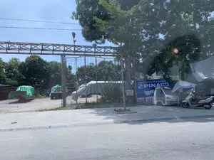 Bức ảnh chụp một quang cảnh ven đường vào một ngày nắng. Ở phía trước là mặt đường nhựa và vỉa hè. Phía sau vỉa hè là một khu đất được rào lại bằng cổng sắt và hàng rào lưới. Bên trong khu đất này, có một vài chiếc xe tải đang đỗ, một số xe được phủ bạt màu xanh hoặc bạt sọc. Đây có vẻ là một bãi đỗ xe hoặc kho chứa hàng của một công ty. Trên vỉa hè, một chiếc xe máy tay ga và một chiếc xe tải nhỏ màu trắng đang đậu dưới bóng cây. Có một chiếc ô che nắng màu xanh trắng được dựng gần đó. Có thể đọc được một số thông tin từ các biển hiệu: 1. Một tấm biển quảng cáo lớn màu xanh ghi: "CÔNG TY CỔ PHẦN PIN ẮC QUY MIỀN N...", logo "PINACO", và "ĐIỆN SEN", cho thấy sự hiện diện của một doanh nghiệp liên quan đến ắc quy. 2. Một tấm biển nhỏ hơn treo trên cây ghi "BÁN ĐẤT" kèm theo số điện thoại "ĐT: 0983050385" và "0912698234". Tổng thể, đây là một khung cảnh tĩnh, không có hoạt động của con người đang diễn ra.。（日本語訳）。写真は晴れた日の道端の光景を写しています。手前にはアスファルトの路面と歩道があります。歩道の後ろには、鉄の門と金網のフェンスで囲まれた土地があります。この土地の中には、数台のトラックが駐車しており、一部の車両は青いシートや縞模様のシートで覆われています。ここは企業の駐車場または資材置き場のようです。歩道では、スクーター1台と白い小型トラック1台が木陰に停まっています。近くには青と白の日よけ傘が立てられています。看板からいくつかの情報を読み取ることができます：1. 青い大きな広告看板には、「CÔNG TY CỔ PHẦN PIN ẮC QUY MIỀN N...」、「PINACO」のロゴ、そして「ĐIỆN SEN」と書かれており、バッテリー関連企業の存在を示しています。2. 木に掛けられた小さな看板には「BÁN ĐẤT」と書かれ、電話番号「ĐT: 0983050385」と「0912698234」が添えられています。全体として、これは静的な光景であり、人間の活動は見られません。
