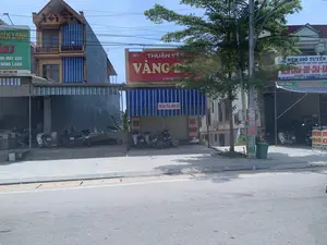 Bối cảnh là một con đường với nhiều cửa hàng kinh doanh san sát nhau tại Việt Nam, cụ thể là trên Quốc lộ 45, Thị trấn Thiệu Hóa, tỉnh Thanh Hóa, dựa trên thông tin từ các biển hiệu. Bầu trời trong xanh, quang đãng. Các cửa hàng là những ngôi nhà ống, vừa để ở vừa để kinh doanh, với nhiều biển hiệu quảng cáo. Có nhiều xe máy đang đậu trên vỉa hè trước các cửa hàng. Khung cảnh khá yên tĩnh, không có hoạt động mua bán rõ rệt.。Nội dung các biển hiệu có thể đọc được:。- Biển hiệu màu xanh lá cây bên trái: "ĐIỆN LẠNH", "...NH-MÁY GIẶT", "...NÓNG LẠNH", "...ẢNH DÀI HẠN", và số điện thoại "...7-0977 890 729".。- Biển hiệu màu đỏ và vàng ở giữa: "THUẬN YÊN VÀNG BẠC", địa chỉ "ĐC: XÓM 3, THIỆ...". Bên dưới là mái hiên sọc xanh trắng có chữ "THUẬN YÊN: NEM GIÒ".。- Biển hiệu bên phải: "NEM GIÒ TUYẾN...", "CHUYÊN SẢN XUẤT KINH DOANH NEM CHUA-GIÒ-CHẢ-BÁNH CHƯNG", "CHẤT LƯỢNG CAO", địa chỉ "ĐC: QL 45-TT THIỆU HÓA-TH" và số điện thoại "ĐT: 0965 248...". Có các biển nhỏ hơn ghi "Tuyến Hằng" và số điện thoại "0965 248 113".。Các vật thể khác trong ảnh bao gồm một cây xanh, một cột điện với hệ thống dây điện chằng chịt, và một thùng rác màu xanh.。（日本語訳）。背景はベトナムのある通りで、看板の情報に基づくと、具体的にはタインホア省ティエウホア町、国道45号線沿いに多くの商店が密集しています。空は青く澄み渡っています。各店舗は住居兼店舗の管状家屋（町家）で、多くの広告看板があります。店の前の歩道には多くのバイクが停まっています。風景はかなり静かで、明確な売買活動は見られません。読み取れる看板の内容：。- 左側の緑色の看板：「ĐIỆN LẠNH（電気・冷凍）」、「...NH-MÁY GIẶT（...洗濯機）」、「...NÓNG LẠNH（...温水・冷水）」、「...ẢNH DÀI HẠN（...長期保証）」、そして電話番号「...7-0977 890 729」。- 中央の赤と黄色の看板：「THUẬN YÊN VÀNG BẠC（トゥアンイエン金銀店）」、住所「ĐC: XÓM 3, THIỆ...（住所：第3組、ティエウ...）」。その下には「THUẬN YÊN: NEM GIÒ（トゥアンイエン：ネム・ヨー）」と書かれた青と白の縞模様の庇があります。- 右側の看板：「NEM GIÒ TUYẾN...（ネム・ヨー・トゥエン...）」、「CHUYÊN SẢN XUẤT KINH DOANH NEM CHUA-GIÒ-CHẢ-BÁNH CHƯNG（ネムチュア、ヨー、チャー、バインチュンの製造販売専門）」、「CHẤT LƯỢNG CAO（高品質）」、住所「ĐC: QL 45-TT THIỆU HÓA-TH（住所：国道45号線-ティエウホア町-タインホア省）」、そして電話番号「ĐT: 0965 248...（電話：0965 248...）」。より小さい看板には「Tuyến Hằng（トゥエン・ハン）」と電話番号「0965 248 113」が書かれています。写真にあるその他の物体には、一本の緑の木、複雑に絡み合った電線のある電柱、そして青いゴミ箱が含まれます。