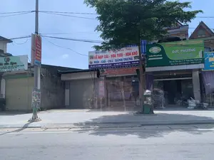 背景はタインホア省ティエウホア町の通りで、商店が隣接して立ち並んでいます。道路と歩道は閑散としており、通行人はいません。昼休みか休日の可能性があります。読み取れる看板の内容：。- 左側の看板：「ハアン・スパ」、専門は「肌の特別治療とケア - ニキビ - シミ - そばかす - クレーター状の傷跡」、「アートメイク - 生徒育成」。住所はティエウホア - TH。この店はシャッターが閉まっています。- 電柱の赤い横断幕：「力を合わせて新しい農村を建設しよう...人民の豊かで幸せな生活のために」と書かれており、VietinBankのロゴがあります。- 中央の大きな看板：「レストラン：キー・チュン・チャン」、専門は「結納品 - 生花 - ドライフラワー」、「ネム - ゾー - チャー - バインチュン」。住所：「タインホア省ティエウホア町第7街区」。この店も閉まっています。- 右側の看板（緑色）：「ドゥック・フオン」店には「ティエンフォン・プラスチック」の看板があり、専門は「電気・水道資材、家庭用電気 - 総合金物」。住所：「タインホア省ティエウホア町第7小区」。この店は開いており、中にはバイク1台と商品の袋があります。写真の中のその他の物体には、多くの電線が絡み合った電柱1本、大きな緑の木1本、そして隣接する建物が含まれます