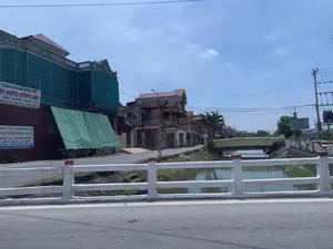 背景はベトナムの町または農村地域にある道で、晴れた日に撮影されたものです。写真は、白いコンクリートの欄干がある橋の上から撮影されており、小さな水路とそれに沿って走る道を見下ろしています。水路の向こう側には、建物や店があります。目立っているのは、「Lâm Hải」（ラム・ハイ）という名前の農産物店の看板がある大きな赤い建物です。これらの看板には、以下の情報が明記されています。- 看板1：「...皮むき緑豆、...農産物、- ゴマ - もち米 - うるち米 - トウモロコシ、...ヒマワリ - 米の春雨 - 海藻の春雨」。- 看板2：「Lâm Hải 各種売買専門、- 醤 - 緑豆 - 皮むき豆 - 落花生 - ゴマ、もち米 - うるち米 - カンナの春雨 - 米の春雨、ヒマワリ - ユウガオの種、...キクラゲ - 乾燥タケノコ - ハスの実。住所：タインホア省ティエウホア町第7地区。電話番号：0914 338 171 - 0395 333 120 - 0964 560 722」。その隣には、緑色の建設用ネットで覆われている別の大きな建物があります。一台の小型トラックが、青と白の縞模様の庇の下、店の前に停まっています。少し先には、水路にかかる別の黄色い橋があります。道を自転車に乗っている人が一人います。その他にも、「H5 14」と書かれた道端のキロポスト、交通標識、電柱、そして入り組んだ電線のような他の物もあります。別の店には、「... RỬA XE」（...洗車）と書かれた看板があります