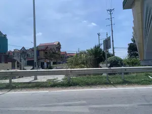 Bức ảnh chụp một khung cảnh ven đường tại một khu vực dân cư ở Việt Nam. Phía trước là một con đường nhựa và một lan can hộ lan bằng kim loại. Phía sau lan can là nhiều ngôi nhà với kiến trúc đa dạng, một số có mái ngói đỏ. Nổi bật trong ảnh là một cổng làng truyền thống, trên đó có dòng chữ 'LÀNG CỔ' và một ký tự Hán Nôm ở giữa, tuy nhiên các chữ khác đã bị mờ. Gần đó có một biển báo giao thông hình tam giác và một biển hiệu quảng cáo dịch vụ 'RỬA XE'. Hệ thống cột điện và dây điện chằng chịt cũng xuất hiện trong khung cảnh. Bầu trời trong xanh, có ít mây.。（日本語訳）。写真はベトナムのある住宅地の道端の風景を撮影したものです。手前にはアスファルトの道路と金属製のガードレールがあります。ガードレールの後ろには、多様な建築様式の家々が多数あり、中には赤い瓦屋根の家もあります。写真の中で目立っているのは伝統的な村の門で、その上には「LÀNG CỔ」という文字と中央に漢字（チュノム）一文字がありますが、他の文字は薄れてしまっています。その近くには三角形の交通標識と、「RỬA XE」（洗車）サービスの広告看板があります。電柱と複雑に絡み合った電線のシステムも風景の中に現れています。空は青く澄んでおり、雲はほとんどありません。