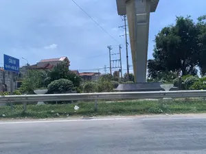 Hình ảnh chụp một khung cảnh ven đường dưới bầu trời xanh. Có một biển báo giao thông màu xanh lam ghi chữ trắng: "CẦU B9" và "Km 61+980 QL.45". Phía trước là một con đường nhựa, tiếp đến là dải cỏ và lan can hộ lan bằng kim loại. Bên phải có một trụ bê tông lớn, có thể là một phần của cầu vượt hoặc đường trên cao. Phía sau lan can là cây cối xanh tươi, bao gồm cây bụi và cây giống cây cọ. Xa hơn là những ngôi nhà dân có mái ngói đỏ, cột điện và đường dây điện. Phía sau biển báo có một cổng làng nhưng chữ viết trên đó không thể đọc rõ.。（日本語訳）。青空の下、道端の風景を撮影した写真です。白い文字で「CẦU B9」および「Km 61+980 QL.45」と書かれた青い交通標識があります。手前にはアスファルトの道路があり、その次に草地と金属製のガードレールがあります。右側には大きなコンクリートの柱があり、陸橋または高架道路の一部である可能性があります。ガードレールの後ろには、低木やヤシのような木々を含む、青々とした木々があります。さらに遠くには、赤い瓦屋根の民家、電柱、そして電線が見えます。標識の後ろには村の門がありますが、そこに書かれている文字ははっきりと読み取れません。