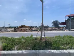 Bối cảnh là một công trường xây dựng nằm cạnh một con đường nhựa vào một ngày nắng. Một chiếc máy xúc màu vàng đang hoạt động, đào đất. Gần đó, một công nhân mặc áo bảo hộ màu cam và đội mũ đang làm việc. Tại công trường có các đống đất, một chồng gạch lát vỉa hè và một vài ống cống. Ở phía sau, có thể thấy các máy móc xây dựng khác như xe tải. Bên phải là một công trình có tường đá và mái tôn màu đỏ. Phía trước là lề đường có cỏ mọc và một cây xanh mới trồng được chống bằng cọc gỗ. Phía xa có những ngọn đồi dưới bầu trời xanh.。（日本語訳）。舞台は晴れた日、アスファルト道路の隣にある建設現場です。一台の黄色いショベルカーが稼働し、土を掘っています。その近くでは、オレンジ色の保護服を着てヘルメットをかぶった作業員が働いています。現場には土の山、積み重ねられた歩道用レンガ、そしていくつかの土管があります。後方には、トラックのような他の建設機械が見えます。右側には、石の壁と赤いトタン屋根の建物があります。手前には草が生えている路肩と、木の杭で支えられた新しく植えられた木があります。遠くには青空の下に丘があります。