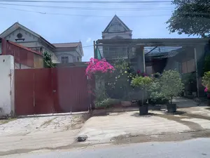 Đây là hình ảnh chụp một con đường trong khu dân cư vào ban ngày. Bối cảnh cho thấy một ngôi nhà có cổng sắt lớn màu đỏ. Trên cột tường trắng bên trái cổng có chữ viết tay, có thể đọc được chữ "VÀO" nhưng các ký tự còn lại không rõ. Bên phải cổng là một khoảng sân có mái che lưới, nơi trồng rất nhiều cây cảnh và hoa trong chậu. Nổi bật là một giàn hoa giấy màu hồng rực rỡ, cùng với các loại hoa màu vàng và nhiều cây xanh khác. Ở phía sau là những ngôi nhà khác với kiến trúc mái ngói. Bầu trời trong xanh và có nhiều đường dây điện chằng chịt ở phía trên. Con đường phía trước là đường bê tông và nhựa đường. Khung cảnh yên tĩnh, không có hoạt động nào của con người diễn ra.。（日本語訳）。これは、昼間の住宅街の道を撮影した写真です。背景には、大きな赤い鉄の門がある家が見えます。門の左側にある白い壁の柱には手書きの文字があり、「VÀO」という文字は読めますが、残りの文字は不明瞭です。門の右側には網の屋根で覆われた庭があり、そこにはたくさんの観葉植物や鉢植えの花が植えられています。鮮やかなピンク色のブーゲンビリアの棚が目立ち、黄色い花や他の多くの緑の植物もあります。後方には、瓦屋根の建築様式の他の家々があります。空は青く澄んでおり、上にはたくさんの電線が張り巡らされています。手前の道はコンクリートとアスファルトの道です。静かな光景で、人の活動は見られません。