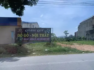 Bức ảnh chụp một khung cảnh ven đường. Có một tấm biển quảng cáo lớn cho một cửa hàng đồ gỗ và nội thất tên là 'Quang Quý'. Nội dung trên biển quảng cáo ghi: 'NỘI THẤT QUANG QUÝ', 'QUANG QUÝ', 'ĐỒ GỖ - NỘI THẤT', 'BÀN GHẾ - GIƯỜNG TỦ - BÀN PHẤN NHẬP KHẨU', 'Đ/C: THÔN 5 - THIỆU TRUNG - THIỆU HOÁ - THANH HOÁ', 'ĐT: 036 357 1686'. Phía sau tấm biển là một khu đất trống có cỏ mọc và một vài cây chuối. Có những ngôi nhà ở hậu cảnh, một con đường nhựa ở phía trước, và nhiều đường dây điện vắt ngang trên bầu trời trong xanh. Ở phía xa có thể thấy hình dáng của một ngọn núi.。（日本語訳）。写真は道端の風景を撮影したものです。「Quang Quý」という名前の木製家具・インテリア店の大きな広告看板があります。看板の内容は、「NỘI THẤT QUANG QUÝ」、「QUANG QUÝ」、「ĐỒ GỖ - NỘI THẤT」、「BÀN GHẾ - GIƯỜNG TỦ - BÀN PHẤN NHẬP KHẨU」、「Đ/C: THÔN 5 - THIỆU TRUNG - THIỆU HOÁ - THANH HOÁ」、「ĐT: 036 357 1686」と書かれています。看板の後ろには、草が生え、数本のバナナの木がある空き地があります。背景には家々があり、手前にはアスファルトの道路があり、青空には多くの電線が横切っています。遠くには山の姿が見えます。