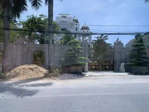 Bối cảnh là một con đường nhựa vào một ngày nắng, nhìn về phía một dinh thự lớn, sang trọng có kiến trúc giống như một tòa lâu đài. Tòa nhà chính được sơn màu trắng, có nhiều tầng với các mái vòm được trang trí hoa văn cầu kỳ. Toàn bộ khu đất được bao quanh bởi một bức tường rào bê tông kiên cố, trên tường có các ô cửa sổ với song sắt mỹ thuật màu vàng. Cổng chính rất lớn và công phu, làm bằng kim loại màu vàng và đen, có các chi tiết trang trí phức tạp và hai tượng sư tử đá ở hai bên. Trong khuôn viên và dọc theo tường rào có trồng nhiều cây xanh, bao gồm cây cọ và các cây cảnh được tạo dáng. Bên ngoài lề đường, có một đống cát và một đống sỏi, cho thấy có thể đang có hoạt động xây dựng hoặc cải tạo. Phía trên không gian là một mạng lưới dây điện và cáp viễn thông chằng chịt. Không có người nào xuất hiện trong ảnh.。（日本語訳）。晴れた日のアスファルトの道が背景で、城のような建築様式の大きくて豪華な邸宅の方を向いている。主な建物は白く塗られており、凝った模様で装飾されたアーチを持つ多層階建てである。敷地全体は頑丈なコンクリートの塀で囲まれており、塀には黄色い装飾的な鉄格子付きの窓がある。正門は非常に大きくて精巧で、黄色と黒の金属でできており、複雑な装飾のディテールと両側に2つの石の獅子像がある。敷地内と塀沿いには多くの緑の木々が植えられており、ヤシの木や形を整えられた観賞用の木々が含まれる。路肩の外側には、砂の山と砂利の山があり、建設または改修作業が行われている可能性を示している。上空には、電線と通信ケーブルが複雑に張り巡らされている。写真には誰も写っていない。