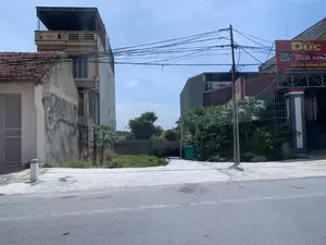 Bức ảnh chụp một con đường nhựa trong một khu dân cư ở Việt Nam. Bên phải đường là một cửa hàng có biển hiệu lớn màu đỏ. Trên biển hiệu có ghi tên cửa hàng là 'Đức' và các dịch vụ như 'RỬA KHO...' (chữ không đọc được rõ) và 'ĐÁNH BÓNG KÍNH'. Địa chỉ được ghi là 'ĐC: PHÚC TRIỀN 1 - ĐÔNG THANH - Đ...'. Bên trái là một ngôi nhà cấp bốn cũ có mái ngói đỏ, phía sau là một tòa nhà nhiều tầng. Giữa các tòa nhà là một khu đất trống mọc nhiều cây cối và cỏ dại. Một cột điện đứng bên đường với mạng lưới dây điện chằng chịt vắt ngang qua. Khung cảnh yên tĩnh, không có người qua lại.。（日本語訳）。この写真はベトナムのある住宅街のアスファルト道路を写したものです。道路の右側には、大きな赤い看板を掲げた店があります。看板には店名「Đức」と、「RỬA KHO...」（文字がはっきりと読み取れない）や「ĐÁNH BÓNG KÍNH」（ガラス研磨）といったサービスが書かれています。住所は「ĐC: PHÚC TRIỀN 1 - ĐÔNG THANH - Đ...」と記されています。左側には赤い瓦屋根の古い平屋があり、その後ろには多層階の建物があります。建物の間には、多くの木々や雑草が生い茂る空き地があります。道路脇には電柱が立っており、複雑に絡み合った電線が横切っています。静かな光景で、通行人はいません。