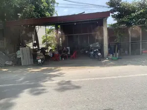 Bức ảnh chụp một cảnh ven đường vào một ngày nắng. Địa điểm dường như là ở một vùng nông thôn hoặc ngoại ô. Có một ngôi nhà hoặc một gian hàng có kết cấu đơn giản với mái che bằng tôn màu đỏ và không gian mở ra đường. Một nhóm ba người đàn ông đang ngồi tụ tập quanh một chiếc bàn nhựa nhỏ màu đỏ, có vẻ như đang thư giãn và trò chuyện. Họ ngồi trên những chiếc ghế nhựa màu đỏ. Một số xe máy được dựng gần đó, cả bên ngoài và bên trong gian nhà. Bên trái, có các tấm lợp fibro xi măng xếp chồng lên nhau. Phía sau nhóm người, không gian có vẻ lộn xộn với nhiều đồ vật khác nhau. Phía trên là cây cối xanh tươi và các đường dây điện. Con đường phía trước được trải nhựa. Không có chữ viết hay biển hiệu nào có thể đọc được.。（日本語訳）。写真は晴れた日の道端の風景を写しています。場所は田舎か郊外のようです。赤いトタン屋根で、道路に面して開かれた空間を持つ、簡素な造りの家か売店があります。3人の男性のグループが、小さな赤いプラスチックのテーブルの周りに集まって座っており、リラックスしておしゃべりをしているようです。彼らは赤いプラスチックの椅子に座っています。近くには、売店の外と中の両方に数台のバイクが停められています。左側には、セメントファイバーの屋根材が積み重ねられています。人々のグループの後ろは、様々な物で散らかっているように見えます。上には青々とした木々と電線があります。前の道は舗装されています。読み取れる文字や看板はありません。