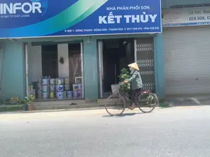 Trên các biển hiệu có ghi:。1. Biển hiệu lớn màu xanh: "INFOR. VẺ ĐẸP TỪ CHẤT LƯỢNG. NHÀ PHÂN PHỐI SƠN KẾT THỦY. ĐỘI 1 - ĐÔNG THANH - ĐÔNG SƠN - THANH HÓA. 0911 258 556 - 0966 538 525".。2. Biển hiệu bên phải, một phần bị che khuất: "AN THUY. Chuyên: CƠ KHÍ, MÁI..., XEN HOA, C...".。Bối cảnh là một con đường nhựa phía trước một dãy cửa hàng. Một người đội nón lá và đeo khẩu trang đang đi xe đạp trên đường. Người này đi ngang qua một cửa hàng đang mở cửa, đây là một nhà phân phối sơn tên là "KẾT THỦY". Bên trong cửa hàng, có thể thấy nhiều thùng sơn được xếp chồng lên nhau. Bên cạnh cửa hàng sơn là một cửa hàng khác đã đóng cửa cuốn, dường như kinh doanh về cơ khí.。（日本語訳）。看板には次のように書かれています：。1. 青い大きな看板：「INFOR. 品質から生まれる美しさ。KET THUY塗料販売店。チーム1 - ドンタイン - ドンソン - タインホア。0911 258 556 - 0966 538 525」。2. 右側の看板、一部が隠れている：「AN THUY。専門：機械、屋根...、花飾り格子、C...」。背景は、店舗が立ち並ぶ前のアスファルトの道路です。菅笠（ノンラー）をかぶり、マスクをつけた人が、道路を自転車で走っています。この人は、開店している店の前を通り過ぎています。これは「KET THUY」という名前の塗料販売店です。店内には、多くの塗料の缶が積み重ねられているのが見えます。塗料店の隣には、シャッターが閉まっている別の店があり、機械関係の事業を営んでいるようです。