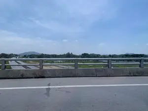 Bức ảnh được chụp từ trên một cây cầu hoặc cầu vượt, nhìn xuống một con đường cao tốc. Ở phía trước là lan can cầu bằng bê tông và kim loại. Bên dưới là đường cao tốc có nhiều làn xe với các phương tiện như ô tô và xe tải đang lưu thông. Xung quanh là khung cảnh nông thôn với những cánh đồng xanh, một số khu vực có vẻ như là ruộng nước hoặc ao hồ. Ở phía xa là những dãy núi và cây cối. Bầu trời trong xanh, có vài gợn mây trắng. Không có biển báo hay chữ viết nào có thể nhìn thấy rõ trong ảnh.。（日本語訳）。写真は橋または高架橋の上から撮影され、高速道路を見下ろしています。手前には、コンクリートと金属でできた橋の欄干があります。下には、乗用車やトラックなどの車両が通行している多車線の高速道路があります。周囲は田園風景で、緑の畑が広がり、一部の地域は水田か池や湖のように見えます。遠くには山並みと木々があります。空は青く澄んでおり、白い雲がいくつか浮かんでいます。