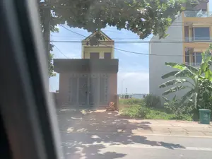 Bức ảnh chụp cảnh quan đường phố vào một ngày nắng, nhìn từ bên trong một phương tiện đang di chuyển. Ở phía bên kia đường là một ngôi nhà có cổng sắt lớn, màu xám, được trang trí hoa văn với một biểu tượng hình tròn ở giữa. Phía sau và bên cạnh là các ngôi nhà cao tầng khác, một số được sơn màu vàng. Cây cối xuất hiện trong ảnh bao gồm một cây lớn có tán lá che phủ một phần phía trên và một bụi chuối ở bên phải. Một thùng rác công cộng màu xanh lá cây được đặt trên vỉa hè. Bầu trời trong xanh, có nhiều đường dây điện giăng qua. Ở phía xa, giữa các ngôi nhà, có thể nhìn thấy một khu vực giống như công trường với các công trình tạm.。（日本語訳）。晴れた日の街の風景を、移動中の乗り物の中から撮影した写真です。道路の向こう側には、中央に円形のシンボルがあしらわれた模様で装飾された、大きな灰色の鉄の門がある家があります。その後ろと隣には他の高層住宅があり、いくつかは黄色に塗られています。写真に写っている木々には、上部の一部を覆う葉冠を持つ大きな木と、右側にあるバナナの茂みが含まれます。緑色の公共ゴミ箱が歩道に置かれています。空は青く澄んでおり、多くの電線が張り巡らされています。遠くの家々の間には、仮設の建造物がある工事現場のような場所が見えます。