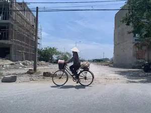 Bối cảnh là một con đường nhựa vào một ngày nắng, hai bên đường là khu vực đang trong quá trình xây dựng. Một người phụ nữ đội nón lá, mặc bộ đồ hoa văn, đang đạp một chiếc xe đạp cũ. Phía trước xe có một cái giỏ đựng túi hàng, và phía sau xe cũng chở một bao tải lớn. Cảnh này cho thấy người phụ nữ có thể đang đi chợ về hoặc đang vận chuyển hàng hóa. Bên trái hình ảnh là một tòa nhà nhiều tầng đang được xây dựng, với kết cấu bê tông và tường gạch còn dang dở, được bao quanh bởi giàn giáo làm bằng tre. Có thể thấy một công nhân đang làm việc trên giàn giáo. Bên phải là một bức tường của một ngôi nhà khác. Con đường còn khá mới nhưng hai bên lề vẫn còn là đất đá và vật liệu xây dựng ngổn ngang. Bầu trời trong xanh, có ít mây. Có các cột điện và dây điện chằng chịt phía trên.。（日本語訳）。背景は晴れた日のアスファルトの道で、道の両側は建設中のエリアです。ノンラーをかぶり、花柄の服を着た一人の女性が、古い自転車をこいでいます。自転車の前には荷物袋が入ったかごがあり、後ろにも大きな袋を積んでいます。この光景は、その女性が市場からの帰り道か、あるいは商品を運んでいるところであることを示唆しています。画像の左側には建設中の多層階の建物があり、コンクリートの構造と未完成のレンガの壁が、竹製の足場で囲まれています。足場の上で作業している一人の作業員が見えます。右側には別の家の壁があります。道はかなり新しいですが、両側の路肩にはまだ土や石、建設資材が散乱しています。空は青く澄んでおり、雲はほとんどありません。上方には電柱と電線が張り巡らされています。