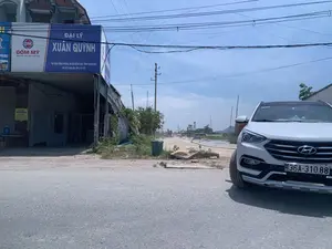 Bối cảnh là một con đường vào một ngày nắng. Bên trái là một tòa nhà một tầng, có vẻ là một đại lý vật liệu xây dựng với các biển hiệu lớn. Một biển hiệu màu xanh và trắng ghi 'ĐẠI LÝ XUÂN QUỲNH', địa chỉ 'THỊ TRẤN RỪNG THÔNG, HUYỆN ĐÔNG SƠN, TỈNH THANH HOÁ' và số điện thoại 'ĐT: 0913 040 635, 0916 771 977'. Ngay bên cạnh là biển hiệu 'GỐM MỸ' với logo 'GM' và khẩu hiệu 'Đẹp nơi nơi, bền mãi mãi'. Phía trước là một con đường nhựa, và một chiếc ô tô SUV màu trắng đang đỗ ở lề đường bên phải. Nhìn sâu vào trong là một con đường đất, có vẻ đang trong quá trình thi công với máy móc xây dựng ở phía xa. Khung cảnh có nhiều cột điện và hệ thống dây điện chằng chịt trên bầu trời xanh. Có một thùng rác màu xanh và cây cỏ mọc ven đường. Bên trong hiên của đại lý, có một tấm biển nhỏ màu vàng ghi 'LIÊN HỆ MUA HÀNG' và một con chó đang nằm nghỉ.。（日本語訳）。背景は晴れた日の道路です。左側には平屋の建物があり、大きな看板から建材代理店のようです。青と白の看板には「ĐẠI LÝ XUÂN QUỲNH」、住所「THỊ TRẤN RỪNG THÔNG, HUYỆN ĐÔNG SƠN, TỈNH THANH HOÁ」、電話番号「ĐT: 0913 040 635, 0916 771 977」と書かれています。そのすぐ隣には、「GM」のロゴと「Đẹp nơi nơi, bền mãi mãi」（どこでも美しく、永遠に長持ち）というスローガンが書かれた「GỐM MỸ」の看板があります。手前にはアスファルトの道路があり、右側の路肩には白いSUV車が駐車しています。奥を見ると未舗装の道路があり、遠くには建設機械が見え、工事中のようです。風景には多くの電柱と、青空に張り巡らされた複雑な電線システムがあります。青いゴミ箱が一つあり、道端には草木が生えています。代理店の軒下には、「LIÊN HỆ MUA HÀNG」（購入連絡先）と書かれた黄色い小さな看板があり、一匹の犬が横になって休んでいます。