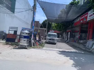 Bối cảnh là một con đường nhỏ được trải bê tông tại một khu vực có nhiều cửa hàng kinh doanh vật liệu xây dựng ở Việt Nam. Ở giữa đường có một chiếc xe tải nhẹ màu trắng hiệu Suzuki đang đỗ, trên thân xe có ghi 'GẠCH MEN ĐỊNH BẮC'. Bên phải là một cửa hàng có mặt tiền màu đỏ, chuyên về vật tư điện nước, với các biển hiệu 'ARISTON' và 'ĐỊNH...'. Địa chỉ trên biển hiệu ghi 'KHU PHỐ THỐNG NHẤT, TT. ĐỒNG THÔNG' và số điện thoại. Phía trước cửa hàng này có trưng bày các phiến đá hoa cương. Bên trái đường, có một bồn nước lớn bằng inox mang nhãn hiệu có chữ 'HA' và các kệ trưng bày các loại ngói lợp. Các biển quảng cáo ở đây có tên thương hiệu 'KATOCO' với dòng chữ 'NGÔI MÁI CÔNG NGHỆ NHẬT BẢN' và 'Amanda'. Phía trên con đường được che bằng một tấm lưới lớn màu đen để che nắng. Trong phông nền, có thể thấy một công trình đang xây dựng và một biển hiệu 'QUÁN NGHĨA HỒNG' và 'BIA VIỆT'.。（日本語訳）。背景は、ベトナムの建材店が多く集まる地域にある、コンクリートで舗装された小さな道です。道の中央にはスズキ製の白い軽トラックが停まっており、車体には「GẠCH MEN ĐỊNH BẮC」と書かれています。右側には、正面が赤い店があり、電気・水道資材を専門としています。看板には「ARISTON」と「ĐỊNH...」と書かれています。看板に書かれた住所は「KHU PHỐ THỐNG NHẤT, TT. ĐỒNG THÔNG」で、電話番号も記載されています。この店の前には、花崗岩の板が展示されています。道の左側には、「HA」という文字のブランドが付いた大きなステンレス製の貯水タンクと、様々な種類の屋根瓦を展示する棚があります。ここの広告看板には、「KATOCO」というブランド名と「NGÔI MÁI CÔNG NGHỆ NHẬT BẢN」（日本の技術による屋根瓦）という文言、そして「Amanda」という名前があります。道路の上は、日差しを遮るために大きな黒い網で覆われています。背景には、建設中の建物と、「QUÁN NGHĨA HỒNG」および「BIA VIỆT」と書かれた看板が見えます。