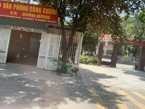 Bức ảnh chụp một con đường vào ban ngày, có nắng và bóng cây râm mát.。Các dòng chữ có thể nhận dạng được bao gồm:。1. Biển hiệu lớn màu đỏ với chữ vàng: "VĂN PHÒNG CÔNG CHỨNG CA ĐÌNH BÌNH".。2. Băng rôn màu đỏ treo ở cổng bên phải, có các khẩu hiệu chính trị, một phần có thể đọc được là "...AN VIỆT NAM QUANG VINH..." và "...ẬP THEO GƯƠNG BÁC HỒ VĨ ĐẠI".。Khung cảnh và hoạt động:。- Địa điểm: Đây là một con đường phố. Bên trái là một tòa nhà là "Văn phòng công chứng", có cổng sắt lớn màu trắng đang mở. Bên trong sân có một vài chiếc xe máy đang đỗ. Bên phải là cổng vào của một cơ quan hoặc trường học, với các cột trụ lớn và hàng rào sắt.。- Hoạt động: Khung cảnh khá yên tĩnh, không có hoạt động mua bán hay giao thông tấp nập. Có người và xe máy bên trong văn phòng công chứng nhưng không rõ hoạt động cụ thể.。- Các vật thể: Tòa nhà, biển hiệu, cổng sắt, xe máy, cây xanh, thùng rác công cộng màu xanh lá cây, vỉa hè, lòng đường có vạch kẻ vàng, cờ đỏ sao vàng (quốc kỳ Việt Nam) và cờ phướn được treo ở cổng cơ quan bên phải.。（日本語訳）。写真は、日中のある道を写しており、日が差し、涼しい木陰があります。認識可能な文字列は以下の通りです：。1. 黄色い文字が書かれた大きな赤い看板：「VĂN PHÒNG CÔNG CHỨNG CA ĐÌNH BÌNH」（カ・ディン・ビン公証役場）。2. 右側の門に掲げられた赤い横断幕には政治的なスローガンがあり、一部読み取れるのは「...AN VIỆT NAM QUANG VINH...」（...栄光あるベトナム...）と「...ẬP THEO GƯƠNG BÁC HỒ VĨ ĐẠI」（...偉大なホーおじさんの模範に倣って...）です。風景と活動：。- 場所：これは街の通りです。左側は「公証役場」の建物で、大きな白い鉄の門が開いています。敷地内には数台のバイクが停まっています。右側は、大きな柱と鉄柵のある官公庁または学校の入り口の門です。- 活動：風景はかなり静かで、活発な売買活動や交通の混雑はありません。公証役場の内部には人とバイクがいますが、具体的な活動は不明です。