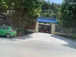 Hình ảnh chụp lối vào trụ sở của một cơ quan nhà nước. Phía trên cổng vào có một tấm biển lớn màu xanh dương với các dòng chữ: 'BẢO HIỂM XÃ HỘI TỈNH THANH HOÁ', 'BẢO HIỂM XÃ HỘI HUYỆN ĐÔNG SƠN', và khẩu hiệu 'BẢO HIỂM Y TẾ - ĐÓNG GÓP KHI LÀNH, ĐỂ DÀNH KHI ỐM'. Đây là trụ sở của Bảo hiểm Xã hội huyện Đông Sơn, tỉnh Thanh Hóa. Bên trái hình ảnh, một chiếc xe taxi màu xanh lá cây đang đỗ bên lề đường. Lối vào sân của trụ sở được lát gạch, bên trong có vài chiếc ô tô đang đỗ. Khung cảnh xung quanh có nhiều cây cối xanh tươi và phía sau là một ngọn đồi.。（日本語訳）。ある国家機関の本部の入り口を撮影した写真。入り口の門の上には、青い大きな看板があり、以下の文字が書かれている：「タインホア省社会保険」、「ドンソン県社会保険」、そしてスローガン「健康保険 - 元気な時に貢献し、病気の時に備える」。これはタインホア省ドンソン県の社会保険事務所の本部である。写真の左側には、緑色のタクシーが道端に停車している。本部の敷地への入り口はタイルで舗装されており、中には数台の車が駐車している。周囲の景色には緑豊かな木々が多く、後ろには丘がある。