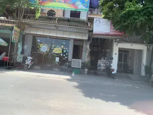 Bối cảnh là một con phố vào ban ngày với nhiều cửa hàng và nhà ở san sát nhau. Ở trung tâm là một trường mẫu giáo có tên "MẦM NON ÁNH DƯƠNG" với mặt tiền bằng kính được trang trí. Tấm biển phía trên ghi rõ địa chỉ: "TT RỪNG THÔNG - HUYỆN ĐÔNG SƠN - TỈNH THANH HÓA" và số điện thoại "ĐT: 0981.228.136". Bên phải là một cửa hàng tạp hóa có biển quảng cáo dầu ăn "MEIZAN GOLD" với dòng chữ "Tuyệt phẩm chiên rán nhiệt độ cao". Biển hiệu của cửa hàng này có nội dung "BIA RƯỢU, BÁ[NH]..." và địa chỉ "ĐC: SN 61, Phố Cao Sơn, TT. Rừng Thông, Đông Sơn, Thanh Hóa". Bên trái là một quán ăn nhỏ có biển hiệu ghi các món "BÚN CHẢ", "BÁNH MỲ CA TE", "XÔI XÉO". Một vài chiếc xe máy đang đậu trên vỉa hè trước các cửa hàng. Cây cối xanh tươi che bóng mát một phần đường và vỉa hè.。（日本語訳）。背景は、多くの店や家が立ち並ぶ昼間の通りです。中央には「MẦM NON ÁNH DƯƠNG」という名前の幼稚園があり、ガラス張りのファサードが装飾されています。上の看板には住所「TT RỪNG THÔNG - HUYỆN ĐÔNG SƠN - TỈNH THANH HÓA」と電話番号「ĐT: 0981.228.136」が明記されています。右側には食料品店があり、「MEIZAN GOLD」という食用油の広告看板には「高温での揚げ物の傑作」というキャッチコピーが書かれています。この店の看板には「BIA RƯỢU, BÁ[NH]...」という内容と、住所「ĐC: SN 61, Phố Cao Sơn, TT. Rừng Thông, Đông Sơn, Thanh Hóa」が書かれています。左側には小さな食堂があり、看板には「BÚN CHẢ」、「BÁNH MỲ CA TE」、「XÔI XÉO」といった料理名が書かれています。数台のバイクが店の前の歩道に停まっています。緑豊かな木々が道路と歩道の一部に日陰を作っています。