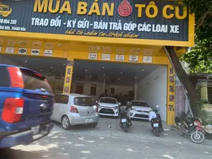 Đây là hình ảnh chụp một cửa hàng kinh doanh ô tô đã qua sử dụng. Biển hiệu lớn phía trên có chữ màu vàng "MUA BÁN Ô TÔ CŨ" và các dịch vụ đi kèm bằng chữ trắng "TRAO ĐỔI - KÝ GỬI - BÁN TRẢ GÓP CÁC LOẠI XE". Bên dưới là khẩu hiệu "chữ tín - niềm tin - trách nhiệm". Trên biển hiệu còn có thông tin địa chỉ "ĐÔNG SƠN - THANH HÓA" và số điện thoại. Dọc hai bên cửa ra vào có biển quảng cáo "MUA XE CŨ GIÁ..." và "HỖ TRỢ TRẢ GÓP 70%". Bên trong và phía trước cửa hàng có một số ô tô và xe máy đang đỗ. Cụ thể, có một chiếc ô tô hatchback màu bạc, hai chiếc sedan màu trắng đang được trưng bày. Một phần của chiếc xe bán tải màu xanh lam đang di chuyển ở tiền cảnh. Trên dầm ngang của biển hiệu có logo của nhiều hãng xe phổ biến như Toyota, KIA, Ford, Suzuki, Mazda, Audi, Hyundai, và Lexus.。（日本語訳）。これは中古車を販売する店の写真です。上部の大きな看板には、黄色の文字で「MUA BÁN Ô TÔ CŨ」と、付随サービスとして白色の文字で「TRAO ĐỔI - KÝ GỬI - BÁN TRẢ GÓP CÁC LOẠI XE」と書かれています。その下には「chữ tín - niềm tin - trách nhiệm」というスローガンがあります。看板には「ĐÔNG SƠN - THANH HÓA」という住所と電話番号の情報もあります。出入り口の両脇には、「MUA XE CŨ GIÁ...」と「HỖ TRỢ TRẢ GÓP 70%」という広告看板があります。店内と店の前には、数台の自動車とバイクが駐車しています。具体的には、銀色のハッチバック車が1台、白色のセダンが2台展示されています。前景では青いピックアップトラックの一部が移動しています。看板の横梁には、トヨタ、KIA、フォード、スズキ、マツダ、アウディ、ヒュンダイ、レクサスといった多くの人気自動車メーカーのロゴがあります。