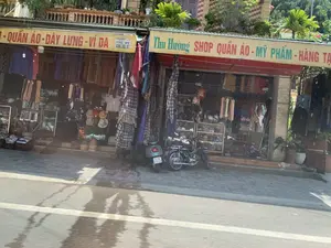 Bức ảnh chụp một con phố với hai cửa hàng bán lẻ nằm cạnh nhau, có vẻ được chụp từ một phương tiện đang di chuyển.。**Thông tin chữ viết:**。- Biển hiệu cửa hàng bên phải: "Thu Hường SHOP QUẦN ÁO - MỸ PHẨM - HÀNG TẠ..." (chữ cuối bị che khuất).。- Biển hiệu cửa hàng bên trái: "...- QUẦN ÁO - DÂY LƯNG - VÍ DA". Bên dưới có một biển nhỏ hơn ghi "PHÚ MỸ" và số điện thoại "0984.103.117".。**Mô tả quang cảnh và hoạt động:**。- **Loại địa điểm:** Đây là một con phố có các cửa hàng kinh doanh, một khu vực buôn bán ở địa phương.。- **Hoạt động cụ thể:** Các cửa hàng đang mở cửa kinh doanh. Hoạt động chính là buôn bán quần áo, phụ kiện thời trang (dây lưng, ví da), mỹ phẩm và hàng tiêu dùng. Hàng hóa được treo và trưng bày dày đặc ở mặt tiền để thu hút khách.。- **Các vật thể trong ảnh:** Nhiều loại quần áo, áo sơ mi, quần dài được treo trên giá. Có các loại mũ, nón được xếp chồng lên nhau trên vỉa hè. Hai chiếc xe máy (xe máy) đang đỗ trên vỉa hè trước cửa hàng. Vỉa hè được lát gạch. Phía trên mái hiên của các cửa hàng là ban công nhà có trồng cây cảnh.。（日本語訳）。隣り合った2つの小売店が並ぶ通りを写した写真で、移動中の乗り物から撮影されたようです。**書かれている情報：**。- 右側の店の看板：「Thu Hường SHOP 衣料品 - 化粧品 - 雑貨...」（最後の文字は隠れている）。- 左側の店の看板：「...- 衣料品 - ベルト - 革財布」。その下には「PHÚ MỸ」と電話番号「0984.103.117」が書かれた小さな看板がある。**風景と活動の説明：**。- **場所の種類：** ここは商店が並ぶ通りで、地元の商業地域です。- **具体的な活動：** 店は営業中です。主な活動は、衣料品、ファッションアクセサリー（ベルト、革財布）、化粧品、消費財の販売です。客を引きつけるために、商品は店先にびっしりと掛けられ、陳列されています。- **写真の中の物体：** 多くの種類の衣類、シャツ、ズボンがラックに掛けられています。歩道には様々な種類の帽子が積み重ねられています。店の前の歩道には2台のバイク（xe máy）が駐車されています。歩道はタイルで舗装されています。店の軒の上には、観葉植物が植えられた家のバルコニーがあります。