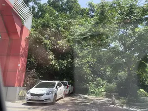 Bức ảnh chụp một khung cảnh ven đường vào ban ngày. Có hai chiếc ô tô màu trắng đang đỗ dưới bóng râm của những tán cây xanh rậm rạp. Chiếc xe ở gần hơn là một chiếc sedan, và phía sau nó là một chiếc xe hatchback. Bên trái là một phần của một công trình kiến trúc lớn, hiện đại, được sơn màu đỏ, với các thanh kim loại màu trắng trang trí ở phía trên. Phần còn lại của khung cảnh được bao phủ bởi cây cối và bụi rậm. Mặt đất là một bãi đỗ xe hoặc vỉa hè. Không có hoạt động nào của con người diễn ra, chỉ có những chiếc xe đang đỗ. Trên cột màu đỏ có một nhãn dán và trên đế bê tông có hình vẽ nhưng không thể đọc được nội dung.。（日本語訳）。写真は、日中の道端の風景を写したものです。緑豊かな木々の木陰に、2台の白い車が駐車しています。手前の車はセダンで、その後ろにはハッチバック車があります。左側には、赤く塗られた大きな現代的な建造物の一部があり、その上部には白い金属製のバーが装飾されています。風景の残りの部分は、木々や茂みに覆われています。地面は駐車場または歩道です。人の活動は見られず、駐車している車だけがあります。赤い柱にはステッカーが貼られており、コンクリートの土台には何か描かれていますが、その内容は読み取れません。