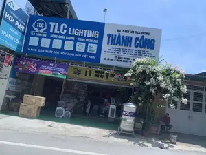 Bức ảnh chụp mặt tiền của một cửa hàng kinh doanh đồ điện máy và vật tư tổng hợp tên là 'Thành Công' vào một ngày nắng. Các biển hiệu lớn cung cấp thông tin chi tiết về các mặt hàng được bán.。Nội dung các biển hiệu:。- Biển hiệu chính: 'ĐẠI LÝ ĐÈN LED TLC LIGHTING THÀNH CÔNG'. Cửa hàng 'CHUYÊN CUNG CẤP: SƠN NỘI THẤT - SƠN NGOẠI THẤT ROSA, THIẾT BỊ ĐIỆN NƯỚC - THIẾT BỊ VỆ SINH - BÌNH NÓNG LẠNH - SEN VÒI, PHỤ KIỆN KIM KHÍ - MÁY LỌC NƯỚC RO THÔNG MINH KAROFI, THIẾT BỊ NHÀ BẾP'. Địa chỉ và số điện thoại: 'ĐC: SỐ 192 - Đ. HOÀNG NGHIÊU - P. ĐÔNG LĨNH', 'ĐT: 0945 85 1992'.。- Biển hiệu bên trái: 'ĐIỆN MÁY GIA DỤNG HÒA PHÁT' và 'ĐIỆN MÁY THÀNH CÔNG'.。- Biển hiệu của hãng đèn: 'TLC LIGHTING - VỮNG CHẤT LƯỢỢNG – TRỌN NIỀM TIN', 'HÃNG SẢN XUẤT ĐÈN LED HÀNG ĐẦU VIỆT NAM'.。- Băng rôn quảng cáo: 'TRỌN VẸN UEFA EURO 2024 TRÊN NỀN TẢNG THỂ THAO SỐ 1' của K+ với chương trình 'GIẢM 50%'.。Cảnh vật và hoạt động:。Đây là một cửa hàng bên đường, đang mở cửa. Phía trước cửa hàng, một người đàn ông mặc áo đỏ đang ngồi trên ghế, bên cạnh một giàn hoa giấy lớn đang nở hoa màu trắng và hồng. Bên trong cửa hàng có một cậu bé. Nhiều sản phẩm được trưng bày bên ngoài, bao gồm một bồn nước inox lớn hiệu 'HXpro', các thùng carton đựng máy điều hòa không khí 'Comfee', và nhiều loại quạt điện được đặt ở lối vào.。（日本語訳）。晴れた日に撮影された、「Thành Công（タインコン）」という名前の電化製品と総合資材を扱う店の正面写真。大きな看板には、販売されている商品に関する詳細な情報が記載されている。看板の内容：。- メイン看板：「TLC LIGHTING LEDランプ代理店 THÀNH CÔNG」。店舗は「専門供給：ROSA室内塗料 - ROSA室外塗料、電気・水道設備 - 衛生設備 - 温水器 - シャワー水栓、金物付属品 - KAROFIスマートRO浄水器、厨房設備」。住所と電話番号：「住所：ドンリン区ホアンギエウ通り192番地」、「電話：0945 85 1992」。- 左側の看板：「HÒA PHÁT（ホアファット）家庭用電化製品」と「THÀNH CÔNG（タインコン）電化製品」。- 照明メーカーの看板：「TLC LIGHTING - 確かな品質、完全な信頼」、「ベトナムのトップLEDランプメーカー」。- 広告バナー：K+の「ナンバー1スポーツプラットフォームでUEFA EURO 2024を完全観戦」と「50%割引」キャンペーン。風景と活動：。これは道端にある店で、営業中である。店の前では、赤いシャツを着た男性が椅子に座っており、その隣には白とピンクの花を咲かせた大きなブーゲンビリアの棚がある。店内には男の子が一人いる。店の外には多くの商品が陳列されており、その中には「HXpro」ブランドの大きなステンレス製貯水タンク、「Comfee」のエアコンが入った段ボール箱、そして入り口に置かれた様々な種類の扇風機が含まれている。