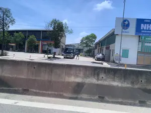 Bức ảnh chụp một khu vực thương mại hoặc công nghiệp ven đường vào một ngày nắng. Ở phía trước là một dải phân cách bằng bê tông, cho thấy ảnh có thể được chụp từ một phương tiện đang di chuyển. Trong sân có một chiếc xe tải nhỏ màu xanh lam đang đỗ với cửa mở, và một người đàn ông đang đứng bên cạnh. Phía sau có một chiếc ô tô khác. Có nhiều tòa nhà với các biển hiệu quảng cáo. Một biển hiệu lớn màu xanh lam ghi 'CON CÒ', 'ACONCO', 'CÔNG TY NH...', 'CHUYÊN KINH DOANH' và địa chỉ 'ĐC: 112 ĐƯỜNG HOÀNG NGHIÊU, P. ĐO...'. Các biển hiệu khác có thể đọc được bao gồm 'PHÂN BÓN PHÚ MỸ Cho mùa bội thu', 'CÔNG TY CỔ PHẦN TẬP ĐOÀN QUỐC TẾ V...', 'Miss COLOR', và 'Ramset'. Một tấm biển viết tay treo trên cây ghi 'CHO THUÊ KHO XƯỞNG'.。（日本語訳）。写真は、晴れた日の道路沿いにある商業または工業地域を撮影したものです。手前にはコンクリート製の中央分離帯があり、写真が移動中の乗り物から撮影された可能性があることを示唆しています。敷地内には青い小型トラックがドアを開けたまま駐車しており、その隣に男性が一人立っています。後ろには別の車があります。多くの建物があり、広告看板が掲げられています。大きな青い看板には、「CON CÒ」、「ACONCO」、「CÔNG TY NH...」、「CHUYÊN KINH DOANH」、そして住所「ĐC: 112 ĐƯỜNG HOÀNG NGHIÊU, P. ĐO...」と書かれています。他に読み取れる看板には、「PHÂN BÓN PHÚ MỸ Cho mùa bội thu」、「CÔNG TY CỔ PHẦN TẬP ĐOÀN QUỐC TẾ V...」、「Miss COLOR」、そして「Ramset」が含まれます。木に掛けられた手書きの看板には、「CHO THUÊ KHO XƯỞNG」と書かれています。