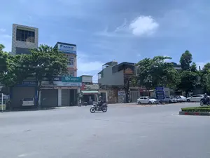 Đây là hình ảnh một góc phố tại một thành phố ở Việt Nam, có thể là ở Thanh Hóa, vào một ngày nắng. Khung cảnh cho thấy một ngã ba đường phố với các hoạt động thường ngày. Một người đàn ông đang lái xe máy trên đường, và một người khác đang đi bộ trên vỉa hè. Nhiều ô tô được đỗ ở lề đường.。Hai bên đường là các tòa nhà nhiều tầng, với nhiều biển hiệu kinh doanh. Các thông tin văn bản có thể đọc được bao gồm:。- Biển tên đường: 'ĐƯỜNG PHÚ THỌ'.。- Biển hiệu của các nhà phân phối thiết bị điện tử và điều hòa không khí: 'DAIKIN - CHUYÊN GIA ĐIỀU HÒA KHÔNG KHÍ HÀNG ĐẦU NHẬT BẢN', 'Panasonic', 'Casper - A BRAND FROM THAILAND'. Các cửa hàng này đều thuộc 'CÔNG TY TNHH SẢN XUẤT & THƯƠNG MẠI ĐÔNG Á - NHÀ PHÂN PHỐI ĐÀO HÒA'.。- Một cửa hàng tên 'H TUẤN THẮNG' tại 'Phú Sơn - Thanh Hóa' với số điện thoại 'ĐT: 0948 909 925 - 0941 260 237'.。- Một quán ăn 'Thanh Thảo CƠM BÌNH DÂN' và 'CƠM BÌNH DÂN - BIA HƠI'.。- Một cụm biển báo màu xanh chỉ dẫn đến 'TRUNG TÂM ĐĂNG KIỂM XE CƠ GIỚI 36-09D', 'TỔNG CÔNG TY ĐÔNG BẮC', và quảng cáo 'CHO THUÊ KHO', 'ÁO DÀI NGỌC HÒA'.。Ngoài ra, có nhiều cây xanh ven đường, bầu trời trong xanh và một số cửa hàng đang đóng cửa cuốn.。（日本語訳）。これは、晴れた日のベトナムのある都市、おそらくタインホアの街角の写真です。日常的な活動が見られる三叉路の光景です。一人の男性が道路でバイクを運転しており、もう一人が歩道を歩いています。多くの車が路肩に駐車されています。道路の両側には多層階の建物が並び、多くの看板が掲げられています。読み取れる文字情報には以下が含まれます：。- 道路名標識：「ĐƯỜNG PHÚ THỌ」。- 電子機器およびエアコンの販売代理店の看板：「DAIKIN - CHUYÊN GIA ĐIỀU HÒA KHÔNG KHÍ HÀNG ĐẦU NHẬT BẢN」、「Panasonic」、「Casper - A BRAND FROM THAILAND」。これらの店舗はすべて「CÔNG TY TNHH SẢN XUẤT & THƯƠNG MẠI ĐÔNG Á - NHÀ PHÂN PHỐI ĐÀO HÒA」に属しています。- 「Phú Sơn - Thanh Hóa」にある「H TUẤN THẮNG」という名前の店で、電話番号は「ĐT: 0948 909 925 - 0941 260 237」です。- 「Thanh Thảo CƠM BÌNH DÂN」と「CƠM BÌNH DÂN - BIA HƠI」という食堂。- 「TRUNG TÂM ĐĂNG KIỂM XE CƠ GIỚI 36-09D」、「TỔNG CÔNG TY ĐÔNG BẮC」への案内、および「CHO THUÊ KHO」、「ÁO DÀI NGỌC HÒA」の広告を示す青い案内標識群。その他、沿道には多くの緑の木々があり、空は青く澄んでおり、いくつかの店はシャッターが閉まっています。