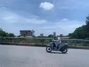Bức ảnh chụp cảnh một người đang lái xe máy trên một cây cầu vào một ngày nắng. Người này mặc bộ đồ chống nắng kín từ đầu đến chân, đội mũ và đeo khẩu trang, một hình ảnh quen thuộc trên đường phố Việt Nam. Chiếc xe máy là loại xe tay ga màu xám. Cây cầu có lan can bằng kim loại. Nhìn qua lan can, có thể thấy khung cảnh của một khu vực đô thị với nhiều cây xanh rậm rạp và các tòa nhà ở phía xa. Trong số các tòa nhà, có một tòa nhà cao tầng với biển hiệu có thể đọc được là 'TRUNG LONG HOTEL'. Bầu trời trong xanh với vài gợn mây, cho thấy thời tiết đẹp.。（日本語訳）。晴れた日に橋の上でバイクを運転している人の光景を写した写真です。この人は頭からつま先までを覆う日焼け防止の服を着て、帽子をかぶり、マスクを着用しており、ベトナムの路上ではおなじみの光景です。バイクは灰色のスクーターです。橋には金属製の手すりがあります。手すり越しには、生い茂る緑樹と遠くの建物がある都市部の風景が見えます。建物の中には、「TRUNG LONG HOTEL」と読み取れる看板のある高層ビルがあります。空は青く澄み、雲がいくつか浮かんでおり、天気が良いことを示しています。