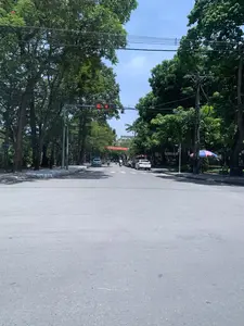 Đây là khung cảnh một con đường đô thị rộng rãi tại Thanh Hóa vào một ngày nắng. Hai bên đường được trồng nhiều cây xanh cổ thụ, tán lá xum xuê tạo bóng mát. Trên đường có một số ô tô và xe máy đang di chuyển và dừng chờ đèn đỏ. Đèn tín hiệu giao thông đang hiển thị số '36' màu đỏ. Một số xe ô tô đang đỗ ở lề đường bên phải. Vắt ngang qua đường có một băng rôn màu đỏ với dòng chữ có thể đọc được một phần là '...PHONG TRÀO THI ĐUA LÀ TRUYỀN THỐNG YÊU NƯỚC CỦA DÂN TỘC TA...'. Ở lề đường bên trái, có một tấm biển nhỏ ghi 'CẮT TÓC'. Trên vỉa hè bên phải, dưới tán cây, có một chiếc ô màu xanh-đỏ và một vài chiếc ghế gỗ.。（日本語訳）。これは晴れた日のタインホア省にある広々とした都市の道路の光景です。道路の両側には多くの古い緑の木が植えられており、青々と茂った葉が木陰を作っています。道路上では、いくつかの自動車とバイクが移動しており、赤信号で停止しています。交通信号機は赤色で「36」という数字を表示しています。いくつかの自動車が右側の路肩に駐車しています。道路を横切って赤い横断幕があり、その一部が「…競争運動は我が民族の愛国の伝統である…」と読める文字が書かれています。左側の路肩には、「散髪」と書かれた小さな看板があります。右側の歩道、木陰の下には、青と赤の傘といくつかの木製の椅子があります。