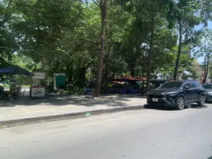 Hình ảnh chụp một khung cảnh ven đường vào ban ngày. Bên phải là một con đường nhựa, có một chiếc ô tô màu đen đang đỗ sát lề. Phần chính của bức ảnh là vỉa hè rộng rãi, rợp bóng cây xanh. Trên vỉa hè có một quán giải khát nhỏ. Có một xe đẩy bán hàng với tấm biển hiệu, trên đó có chữ viết nhưng khó đọc, và hình vẽ một bát đồ ăn. Xung quanh được bố trí bàn ghế và một chiếc ô lớn màu xanh dương để phục vụ khách. Gần đó có một nhà vệ sinh di động màu xanh lá cây. Xa hơn dưới tán cây là nhiều bộ bàn ghế nhựa màu xanh và ô che nắng màu cam. Cảnh vật cho thấy đây là một không gian công cộng, có thể là công viên hoặc một con phố lớn, nơi mọi người có thể ngồi uống nước và thư giãn.。（日本語訳）。昼間の道端の風景を撮影した写真です。右側にはアスファルトの道路があり、黒い自動車が1台、路肩にぴったりと駐車しています。写真の主要部分は、緑の木々に覆われた広々とした歩道です。歩道の上には小さな軽食店があります。看板が付いた屋台があり、そこには文字が書かれていますが読みにくく、一杯の食べ物の絵が描かれています。周りには客をもてなすためのテーブルと椅子、そして大きな青いパラソルが配置されています。その近くには緑色の仮設トイレがあります。さらに奥の木陰には、たくさんの青いプラスチック製のテーブルと椅子、そしてオレンジ色のパラソルがあります。この風景は、ここが公園か大きな通りといった公共の空間であり、人々が座って飲み物を飲んだりリラックスしたりできる場所であることを示しています。