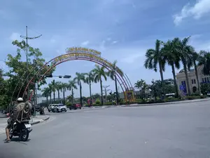 Bức ảnh chụp một góc phố tại một ngã tư hoặc vòng xuyến lớn trong thành phố vào một ngày nắng. Bầu trời trong xanh và có ít mây. Điểm nhấn chính là một cổng chào lớn màu đỏ hình vòng cung bắc qua đường. Trên cổng chào có dòng chữ lớn màu vàng: hàng trên là 'PHỐ ĐI BỘ PHAN CHU TRINH' và hàng dưới là 'VÀ KHÔNG GIAN VĂN HÓA QUẢNG TRƯỜNG LAM SƠN'. Điều này cho thấy đây là lối vào một khu vực công cộng quan trọng. Trên đường có các phương tiện như ô tô và xe máy đang tham gia giao thông. Ở phía trước bên trái, một người đàn ông đội mũ bảo hiểm đang điều khiển xe máy và chở một chiếc xe rùa (xe cút kít) ở phía sau. Hai bên đường và khu vực trung tâm vòng xuyến được trồng nhiều cây xanh, bao gồm cả những hàng cây cọ cao. Phía sau cổng chào là không gian của quảng trường với cây cối và các công trình kiến trúc.。（日本語訳）。写真は、晴れた日の街の大きな交差点またはロータリーの一角を撮影したものです。空は青く澄んでおり、雲はほとんどありません。主な見どころは、道路をまたぐ大きなアーチ型の赤いウェルカムゲートです。ウェルカムゲートには大きな黄色い文字があり、上の行は「PHAN CHU TRINH歩行者天国」、下の行は「およびLAM SON広場文化空間」と書かれています。これは、ここが重要な公共エリアへの入り口であることを示しています。道路には、自動車やバイクなどの車両が通行しています。左手前では、ヘルメットをかぶった男性がバイクを運転し、後ろに手押し車（一輪車）を運んでいます。道路の両側とロータリーの中央エリアには、高いヤシの木を含む多くの緑の木が植えられています。ウェルカムゲートの後ろには、木々や建築物がある広場の空間が広がっています。