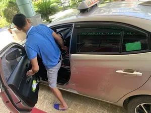 Bức ảnh chụp một người đàn ông đang mở cửa phía người lái của một chiếc xe taxi màu bạc để vào trong. Người đàn ông mặc áo phông polo màu xanh lam, quần soóc màu xám và đi dép lê. Chiếc xe là một chiếc Toyota Vios, có biển hiệu 'TAXI' trên nóc. Trên tấm che mưa ở cửa sổ có chữ 'VIOS'. Trên cửa sổ sau có dán một tem màu xanh lá cây ghi thông tin về mức tiêu thụ nhiên liệu, có thể đọc được các dòng chữ 'MỨC TIÊU THỤ NHIÊN LIỆU', 'Nhãn hiệu: TOYOTA' và các chỉ số '6.1', '8.15', '4.92'. Khung cảnh xung quanh cho thấy đây là khu vực ngoài trời trước một tòa nhà, có sân lát gạch và cây cối trang trí.。（日本語訳）。銀色のタクシーの運転席側のドアを開けて乗り込もうとしている男性の写真です。男性は青いポロシャツ、灰色のショートパンツを着用し、サンダルを履いています。車はトヨタ・ヴィオスで、屋根には「TAXI」の表示灯があります。窓のドアバイザーには「VIOS」の文字があります。後部窓には燃費情報が記載された緑色のステッカーが貼られており、「MỨC TIÊU THỤ NHIÊN LIỆU」、「Nhãn hiệu: TOYOTA」という行、および「6.1」、「8.15」、「4.92」という指標が読み取れます。周囲の風景から、ここが建物の前の屋外エリアで、タイル敷きの中庭と装飾用の植木があることがわかります。