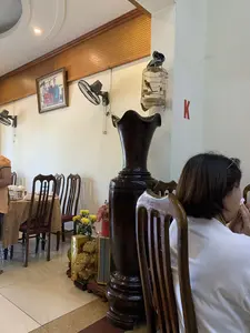 Bối cảnh bên trong một nhà hàng hoặc phòng ăn. Một người phụ nữ mặc áo trắng đang ngồi ở bàn ăn ở phía trước. Không gian được trang trí với bàn ghế gỗ, một bình lục bình lớn bằng gỗ, và các quạt treo tường. Trên tường có treo một lồng chim với một con chim nhỏ bên trong, và một bức tranh đóng khung không rõ nội dung. Trên một cây cột có dán chữ 'K' màu đỏ. Ở phía sau, một bàn ăn khác được trải khăn và bày biện sẵn bát đĩa. Gần đó có các vật trang trí như một chậu hoa cúc vàng và một vật phẩm trang trí màu vàng có thể là cây kim tiền dùng để cầu may mắn.。（日本語訳）。レストランまたは食堂の内部の光景。手前の食卓に、白い服を着た一人の女性が座っている。空間は木製のテーブルと椅子、大きな木製の花瓶、そして壁掛け扇風機で装飾されている。壁には、中に小鳥が一羽いる鳥かごと、内容が不明な額装された絵が一つ掛かっている。一本の柱には、赤い「K」の文字が貼られている。奥には、別の食卓にテーブルクロスが敷かれ、食器が並べられている。その近くには、黄色い菊の鉢植えや、幸運を祈るために使われる金のなる木（キンセンジュ）と思われる黄色い装飾品などの飾り物がある。