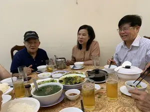 Trên chiếc mũ lưỡi trai màu đen của người đàn ông ngồi bên trái có dòng chữ "Discovery EXPEDITION".。Bối cảnh là một bữa ăn gia đình hoặc một buổi tụ tập thân mật trong nhà. Một nhóm người, bao gồm một người đàn ông lớn tuổi đội mũ, một phụ nữ trung niên và một người đàn ông lớn tuổi đeo kính, đang ngồi quây quần quanh một bàn ăn. Mọi người đang ăn uống và trò chuyện trong không khí ấm cúng.。Trên bàn ăn được bày biện nhiều món ăn Việt Nam. Có thể nhận thấy một tô canh rau lớn, một món xào màu vàng trông giống ốc hoặc lươn, một đĩa lạc rang, cơm trắng, và các bát nước chấm nhỏ có ớt. Ngoài ra còn có một nồi đất màu đen, các cốc bia đá và nước lọc. Mọi người đang sử dụng bát và đũa để thưởng thức bữa ăn.。（日本語訳）。左側に座っている男性の黒い野球帽には、「Discovery EXPEDITION」という文字があります。背景は、家庭での食事、または屋内での親密な集まりです。帽子をかぶった年配の男性、中年の女性、眼鏡をかけた年配の男性を含む一団が、食卓を囲んで座っています。皆は和やかな雰囲気の中で飲食し、会話をしています。食卓には多くのベトナム料理が並べられています。その他にも、黒い土鍋、氷入りのビールのグラス、そして水があります。皆は椀と箸を使って食事を楽しんでいます。