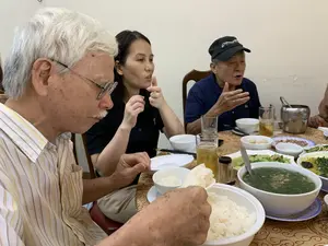 Bức ảnh ghi lại cảnh một bữa ăn gia đình ấm cúng trong nhà. Có ít nhất ba người đang ngồi quanh một bàn ăn. Ở phía bên trái là một ông cụ tóc bạc trắng, đeo kính, mặc áo sơ mi kẻ sọc, đang lấy cơm từ một tô lớn. Ở giữa là một người phụ nữ mặc áo đen đang nói chuyện và ra hiệu bằng tay. Ở phía bên phải là một ông cụ khác đội mũ lưỡi trai màu đen, trên mũ có chữ trắng, có vẻ là logo của "Discovery Channel", ông cũng đang nói chuyện. Trên bàn ăn được trải khăn có hoa văn, bày biện nhiều món ăn đặc trưng của bữa cơm Việt Nam, bao gồm một tô cơm trắng lớn, một tô canh rau, các đĩa thức ăn khác, bát và đũa. Ngoài ra còn có các cốc uống nước, một cốc bia có đá và một xô đá bằng kim loại.。（日本語訳）。この写真は、家の中での温かい家族の食事風景を記録したものです。少なくとも3人が食卓を囲んで座っています。左側には白髪で眼鏡をかけ、ストライプのシャツを着た高齢の男性が、大きな椀からご飯をよそっています。中央には黒い服を着た女性が話しながら手で合図をしています。右側には別の高齢の男性が黒い野球帽をかぶっており、帽子には白い文字で「ディスカバリーチャンネル」のロゴらしきものが見え、彼もまた話しています。模様のあるテーブルクロスが敷かれた食卓には、大きな白米の椀、野菜スープの椀、その他の料理皿、茶碗、箸など、ベトナムの食事に特徴的な多くの料理が並べられています。その他にも、水用のコップ、氷の入ったビールグラス、金属製のアイスペールがあります。
