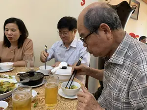 Bức ảnh chụp cảnh một nhóm người đang dùng bữa trong nhà, có thể là tại một nhà hàng. Ở phía trước, một người đàn ông lớn tuổi, tóc đã thưa và bạc, đeo kính và mặc áo sơ mi kẻ caro, đang dùng đũa gắp thức ăn vào bát của mình. Ngồi cạnh ông là một người đàn ông khác đeo kính, mặc áo sơ mi kẻ sọc xanh trắng. Bên trái là một người phụ nữ mặc áo màu nâu. Trên bàn ăn được trải khăn có hoa văn, bày biện nhiều món ăn. Có thể thấy các bát sứ trắng, một nồi đất màu đen, một thố sứ trắng, các ly bia có đá và các đĩa thức ăn, trong đó có một đĩa trông giống món ốc. Trên bức tường phía sau có một chữ cái 'S' màu đỏ, nhưng không có thông tin nào khác có thể đọc được.。（日本語訳）。写真は、屋内で食事をしている人々のグループを写しており、おそらくレストランでの光景です。手前には、髪が薄くなり白髪になった年配の男性が、眼鏡をかけ、チェック柄のシャツを着て、箸で自分の椀に食べ物を取っています。彼の隣には、眼鏡をかけ、青と白のストライプのシャツを着た別の男性が座っています。左側には、茶色い服を着た女性がいます。食卓には模様のあるテーブルクロスが敷かれ、多くの料理が並べられています。白い磁器の椀、黒い土鍋、白い磁器の蓋付きの鉢、氷の入ったビールグラス、そしていくつかの料理皿が見え、その中にはタニシ料理のような一皿があります。後ろの壁には赤い「S」の文字がありますが、他に読み取れる情報はありません。