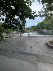 Đây là khung cảnh ven một con sông hoặc kênh đào, có thể là ở thành phố Thanh Hóa. Tiền cảnh là một lối đi bộ rộng rãi được lát gạch hoặc ván, bên cạnh một con đường nhựa. Có nhiều cây xanh được trồng dọc theo lối đi, nổi bật là một cây lớn ở bên trái. Một lan can kim loại chạy dọc theo bờ sông để đảm bảo an toàn. Có một người đang ngồi trên lối đi bộ, cạnh lan can. Phía bên kia sông là một hàng cây rậm rạp và các tòa nhà đô thị. Trên một tòa nhà màu vàng ở phía xa có thể đọc được chữ 'Kanebo'. Bức ảnh hơi mờ, cho thấy nó có thể được chụp từ một phương tiện đang di chuyển.。（日本語訳）。これは川または運河沿いの風景で、おそらくタインホア市のものでしょう。手前には、タイルまたは板で舗装された広い遊歩道があり、その隣にはアスファルトの道路があります。遊歩道に沿って多くの緑の木が植えられており、左側にある大きな木が目立ちます。安全を確保するために、金属製の手すりが川岸に沿って設置されています。遊歩道の上、手すりの隣に座っている人が一人います。川の向こう側には、鬱蒼とした木々の列と都市の建物があります。遠くにある黄色い建物には、「Kanebo」という文字が読み取れます。写真は少しぼやけており、移動中の乗り物から撮影された可能性を示唆しています。